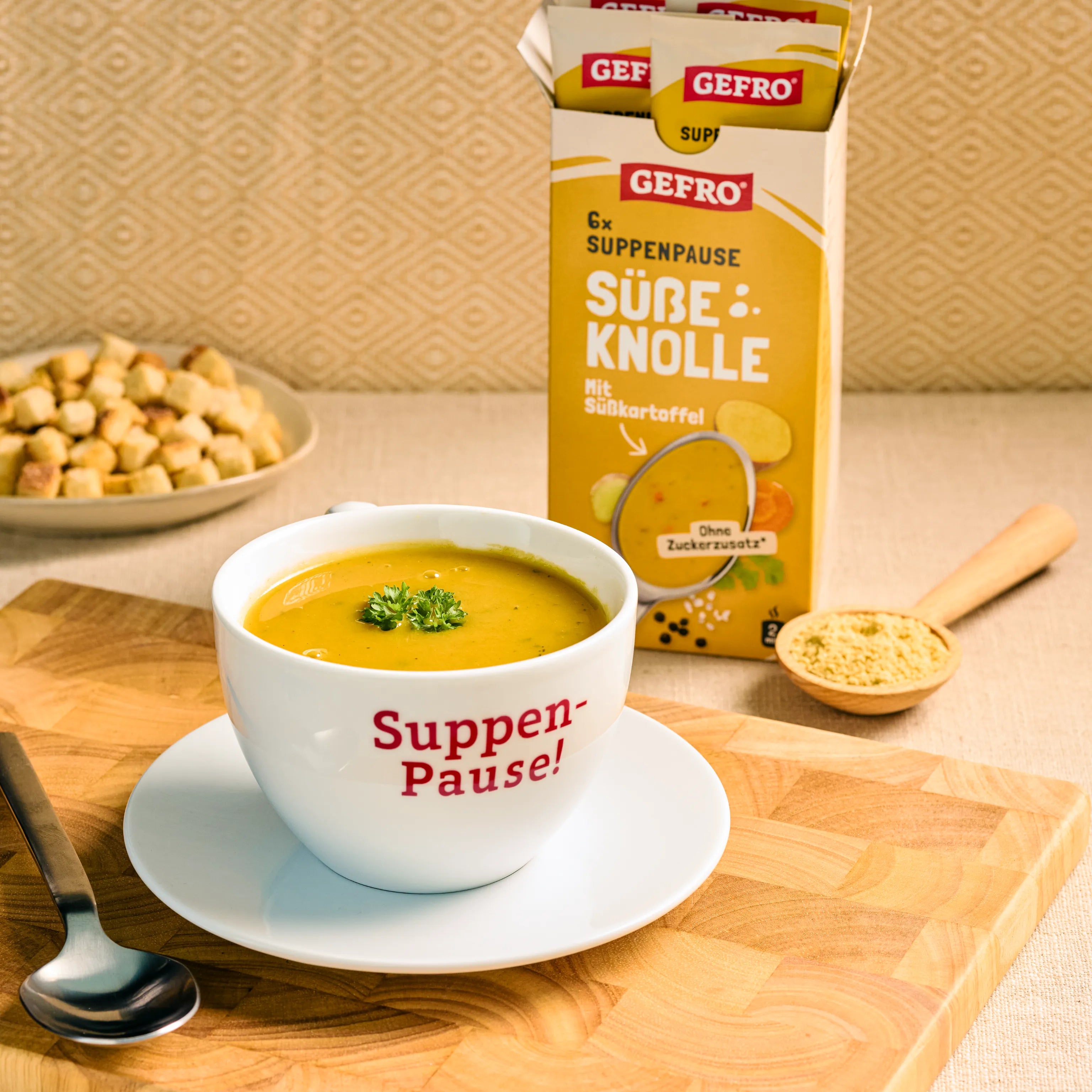 Tasse mit Süßkartoffelsuppe auf Untertasse vor geöffneter GEFRO-Packung „Süße Knolle“ auf Holzbrett mit Löffel