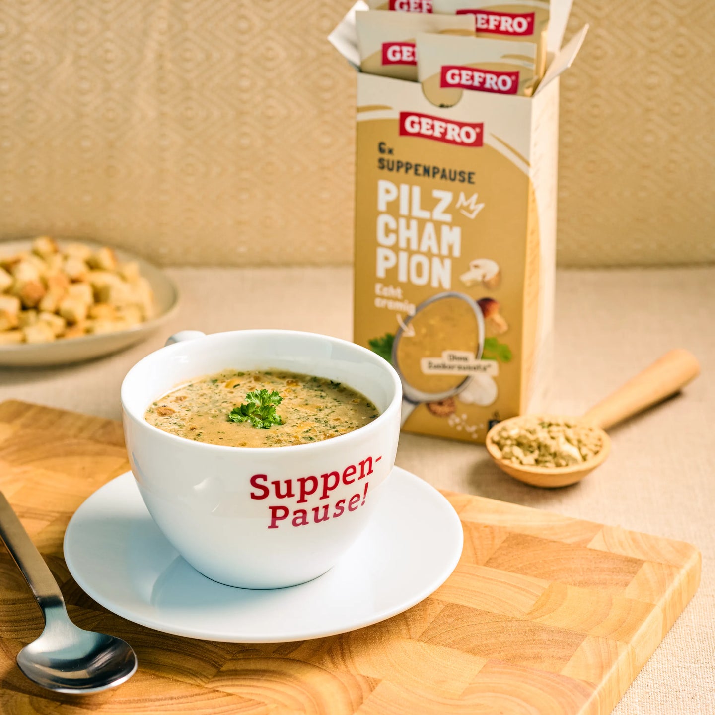 Tasse mit cremiger Pilzsuppe auf Unterteller vor geöffneter GEFRO Verpackung, dazu Löffel und Holzlöffel mit Würze