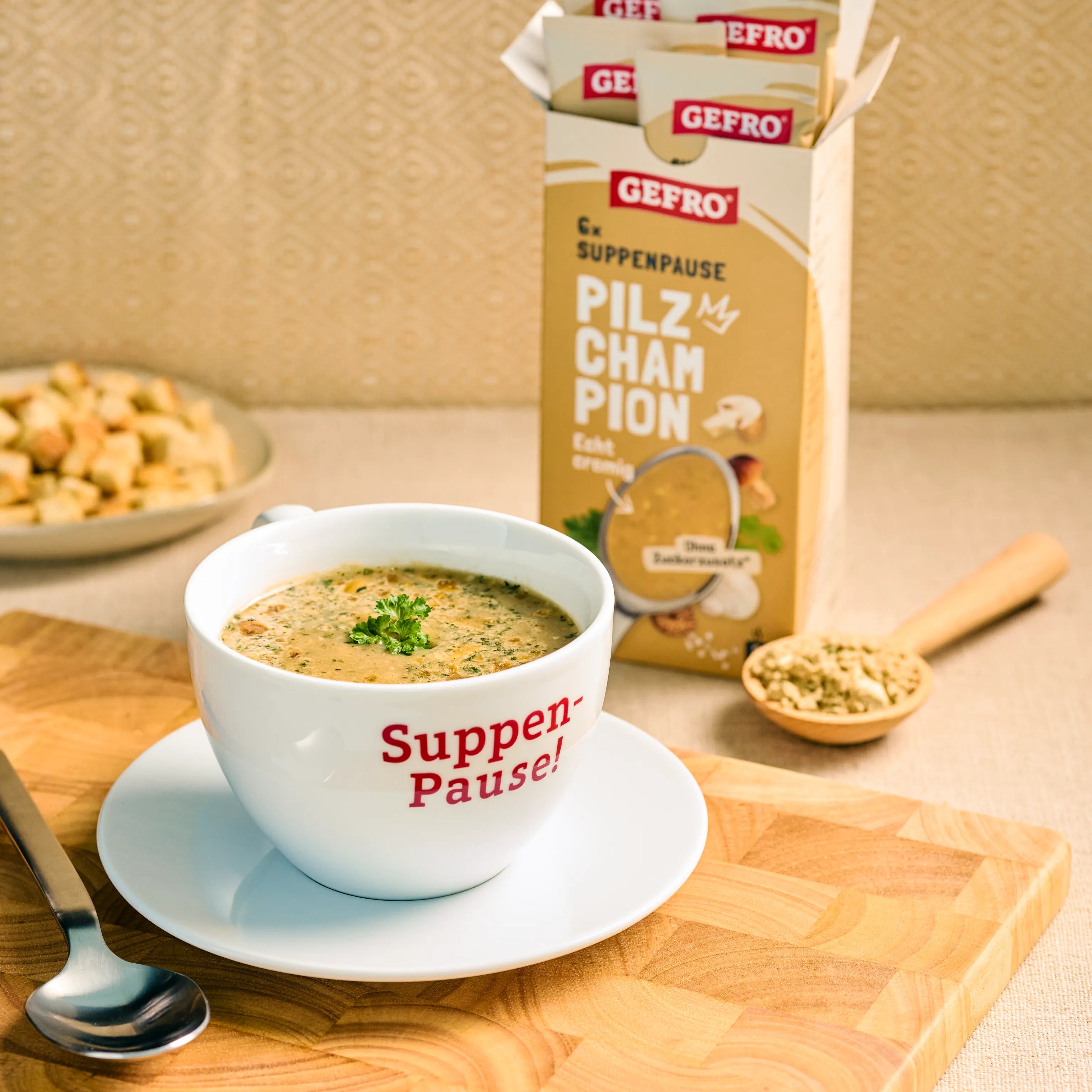 Tasse mit cremiger Pilzsuppe auf Unterteller vor geöffneter GEFRO Verpackung, dazu Löffel und Holzlöffel mit Würze