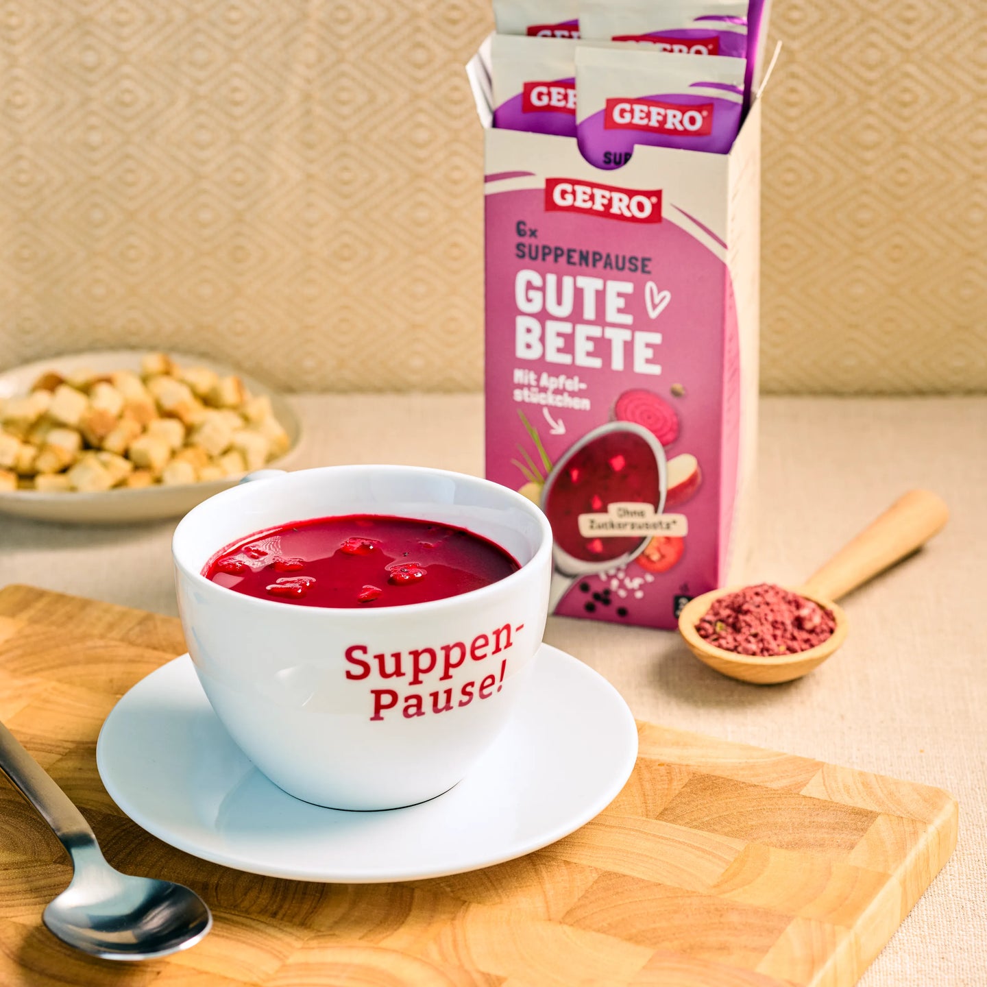 Tasse mit roter Rote-Bete-Suppe vor geöffneter GEFRO-Packung auf Holzbrett, daneben Löffel und Gewürzschale