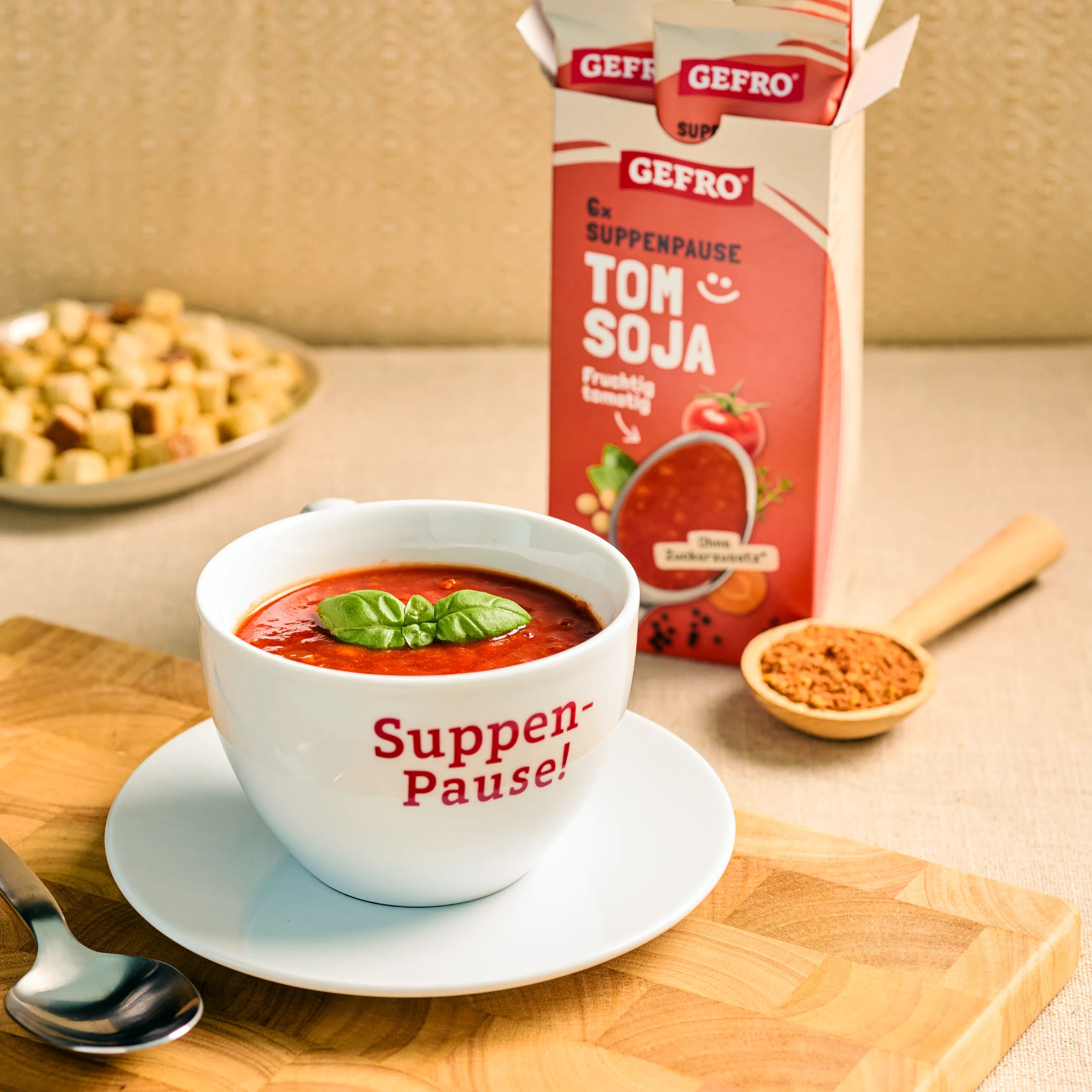 Tasse mit roter Tomaten-Soja-Suppe und Basilikum vor geöffneter GEFRO-Packung auf Holzbrett serviert