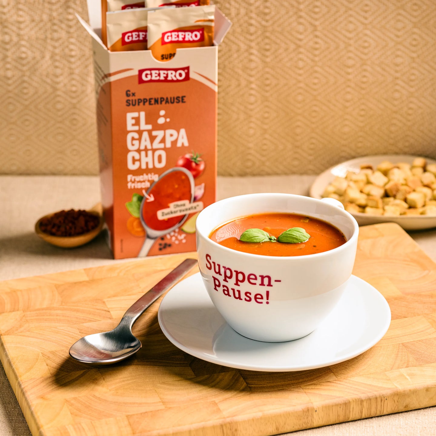 Schale mit roter Gazpacho und Basilikum auf Untertasse, daneben Löffel und geöffnete GEFRO El Gazpacho Packung.