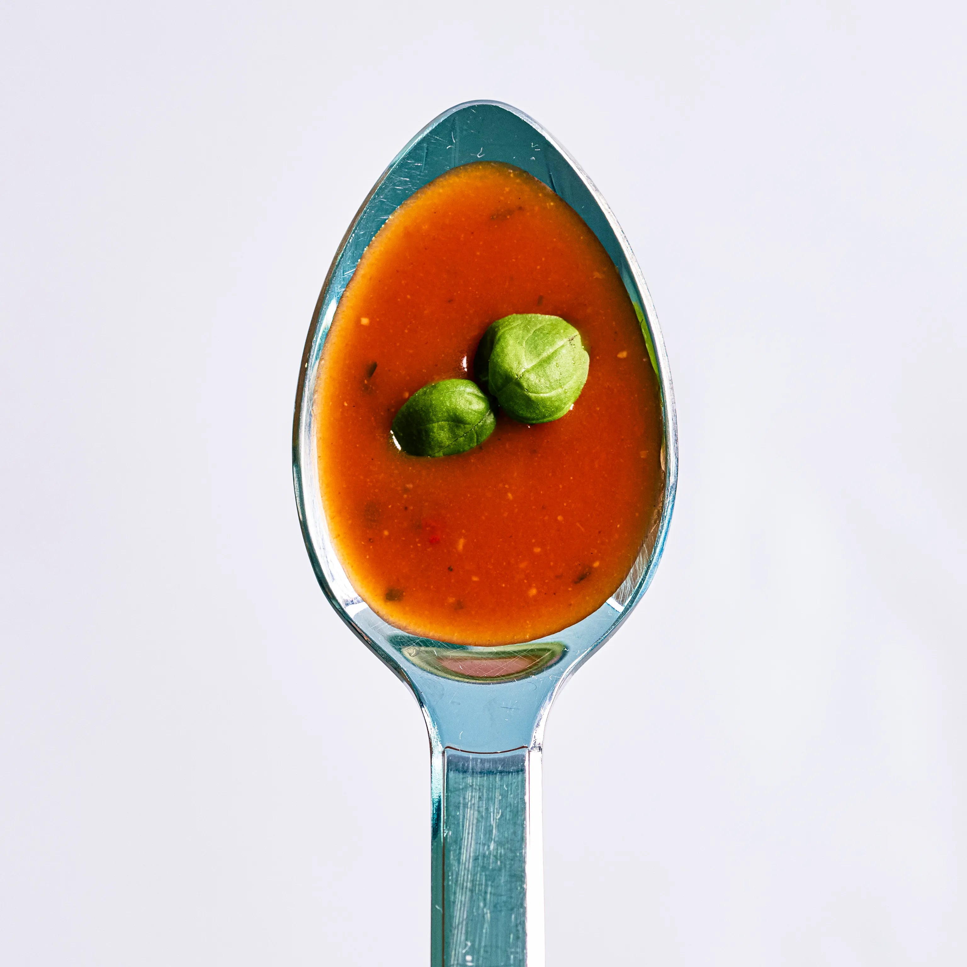 Löffel mit roter Tomatensuppe und zwei grünen Basilikumblättern, Nahaufnahme auf hellem Hintergrund