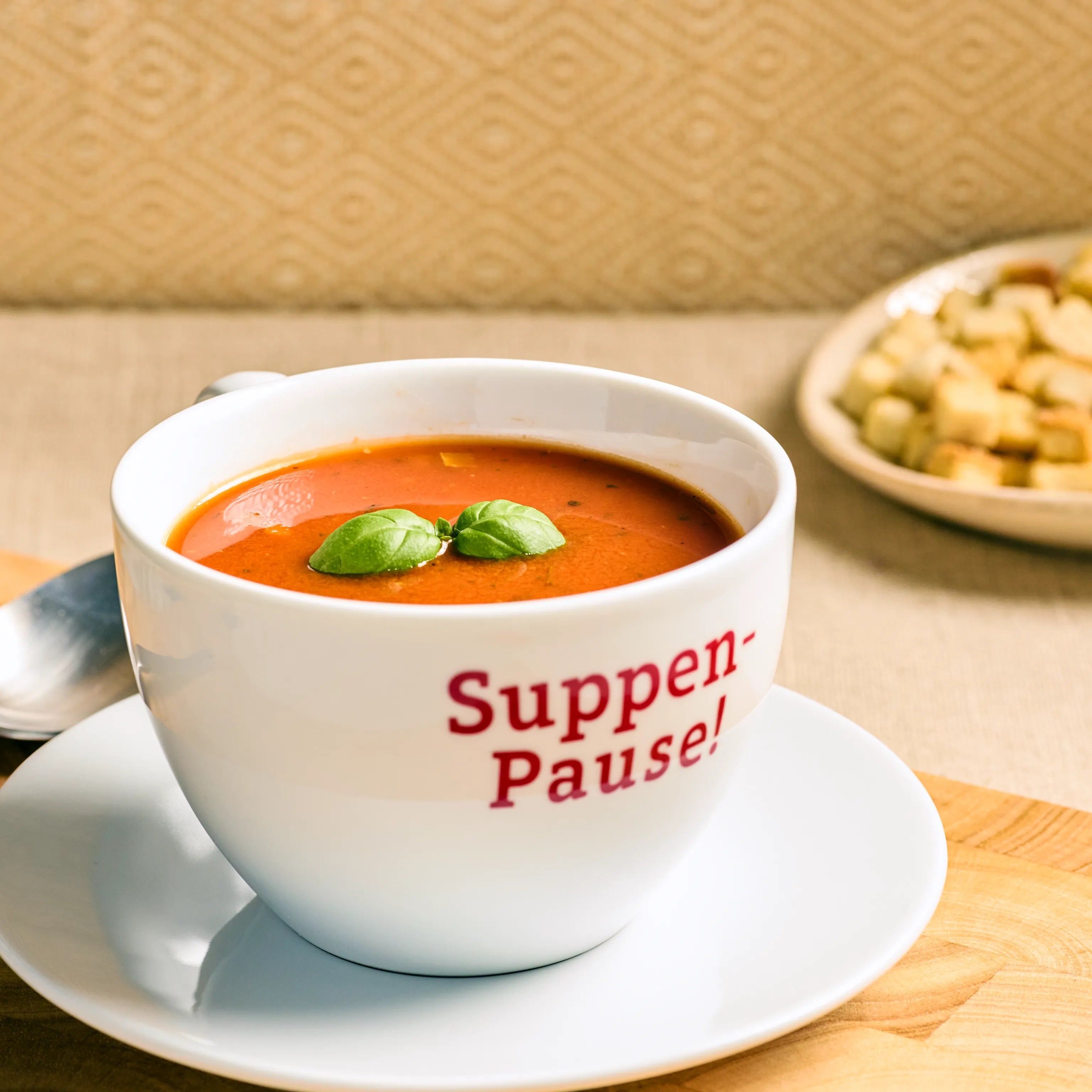 Weiße Tasse mit Tomatensuppe und Basilikum auf Untertasse, dazu Löffel und Brotwürfel im Hintergrund