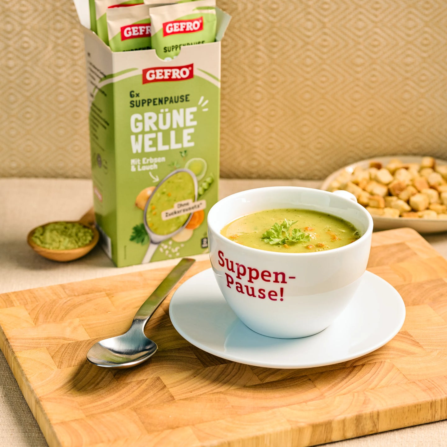 Tasse mit grüner Erbsen-Lauch-Suppe vor GEFRO Verpackung auf Holzbrett, daneben Löffel und Croûtons.