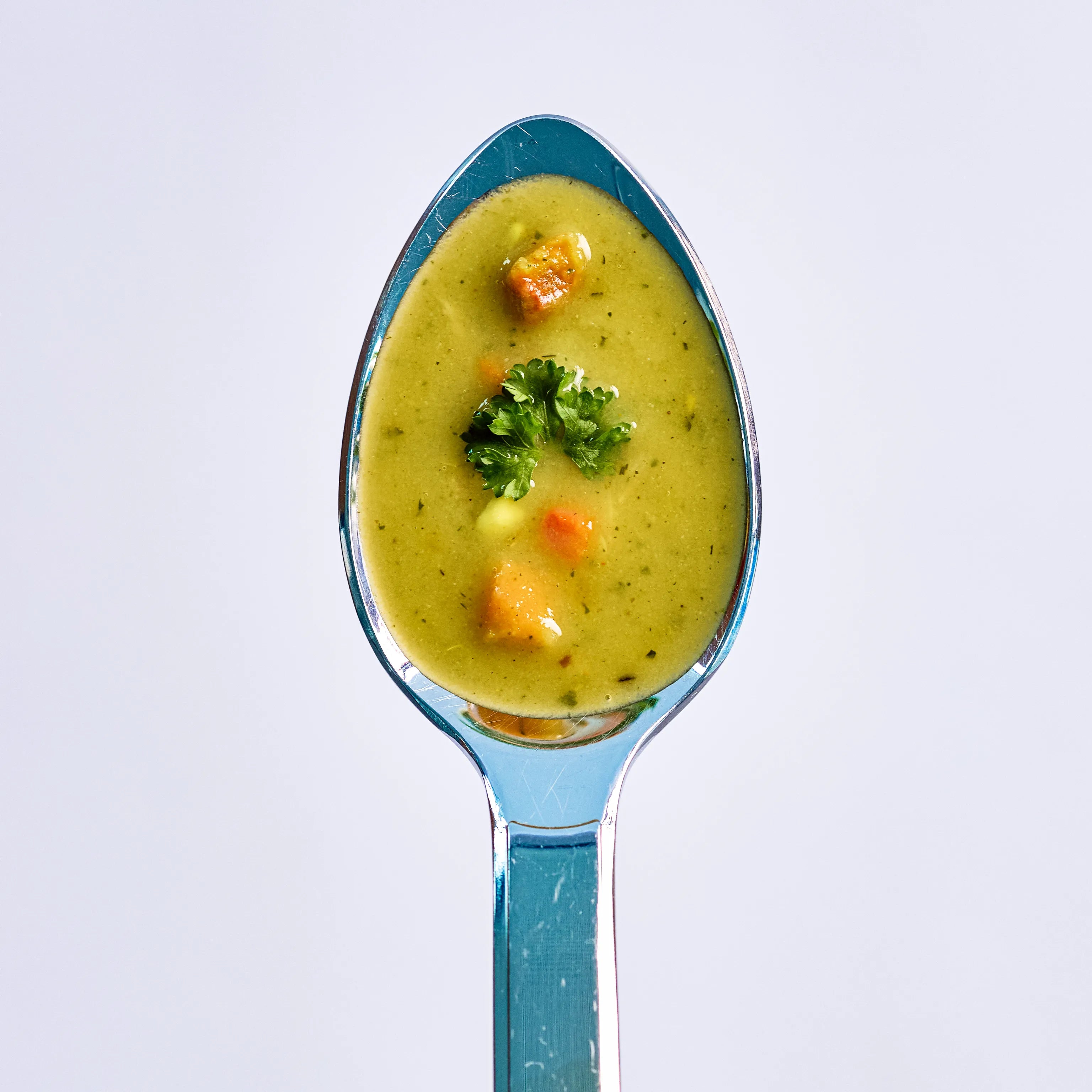 Löffel mit cremiger Gemüsesuppe, garniert mit Petersilie und kleinen Gemüsewürfeln vor hellem Hintergrund