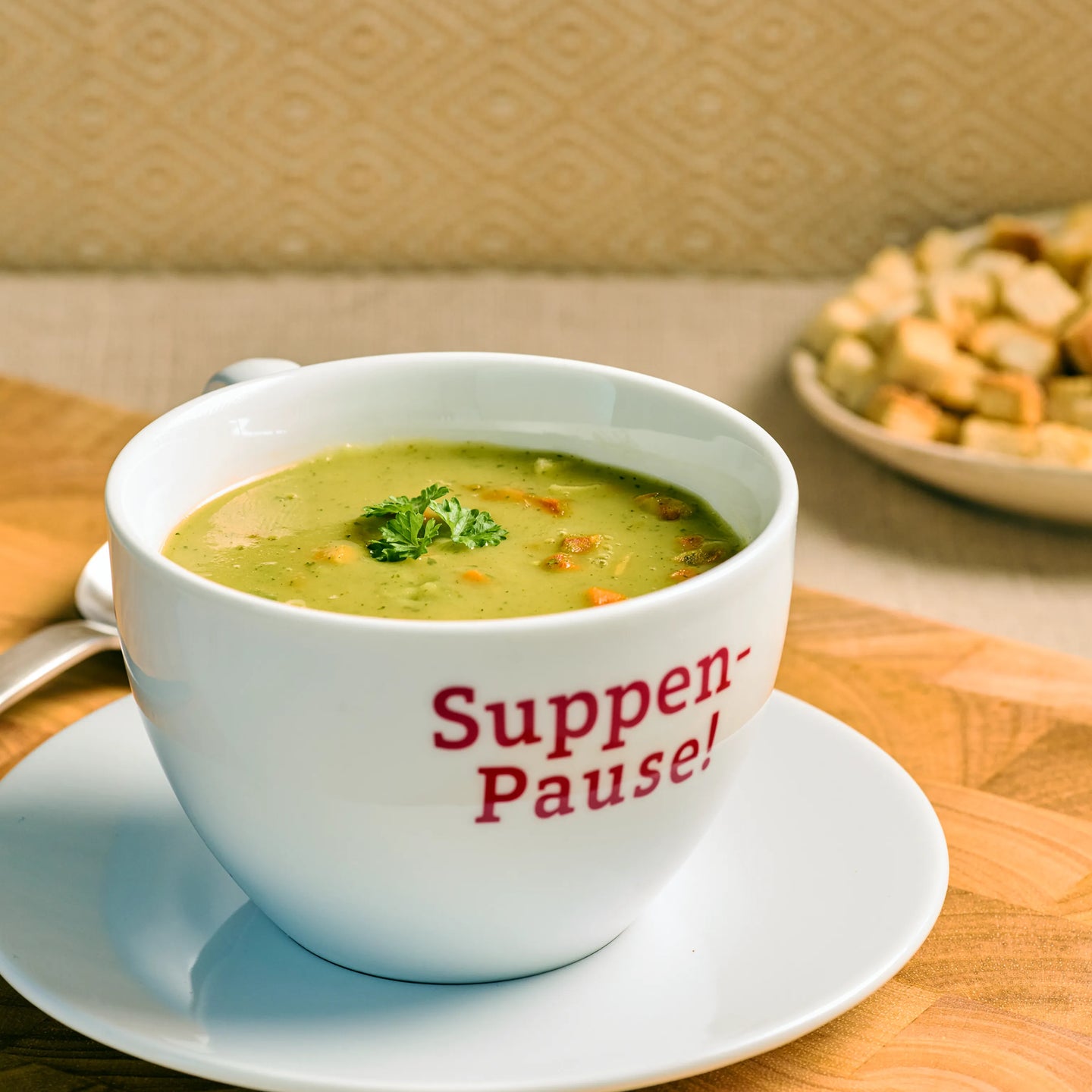 Weiße Tasse mit cremiger Gemüsesuppe auf Unterteller, dekoriert mit Petersilie, daneben Brotwürfel im Hintergrund