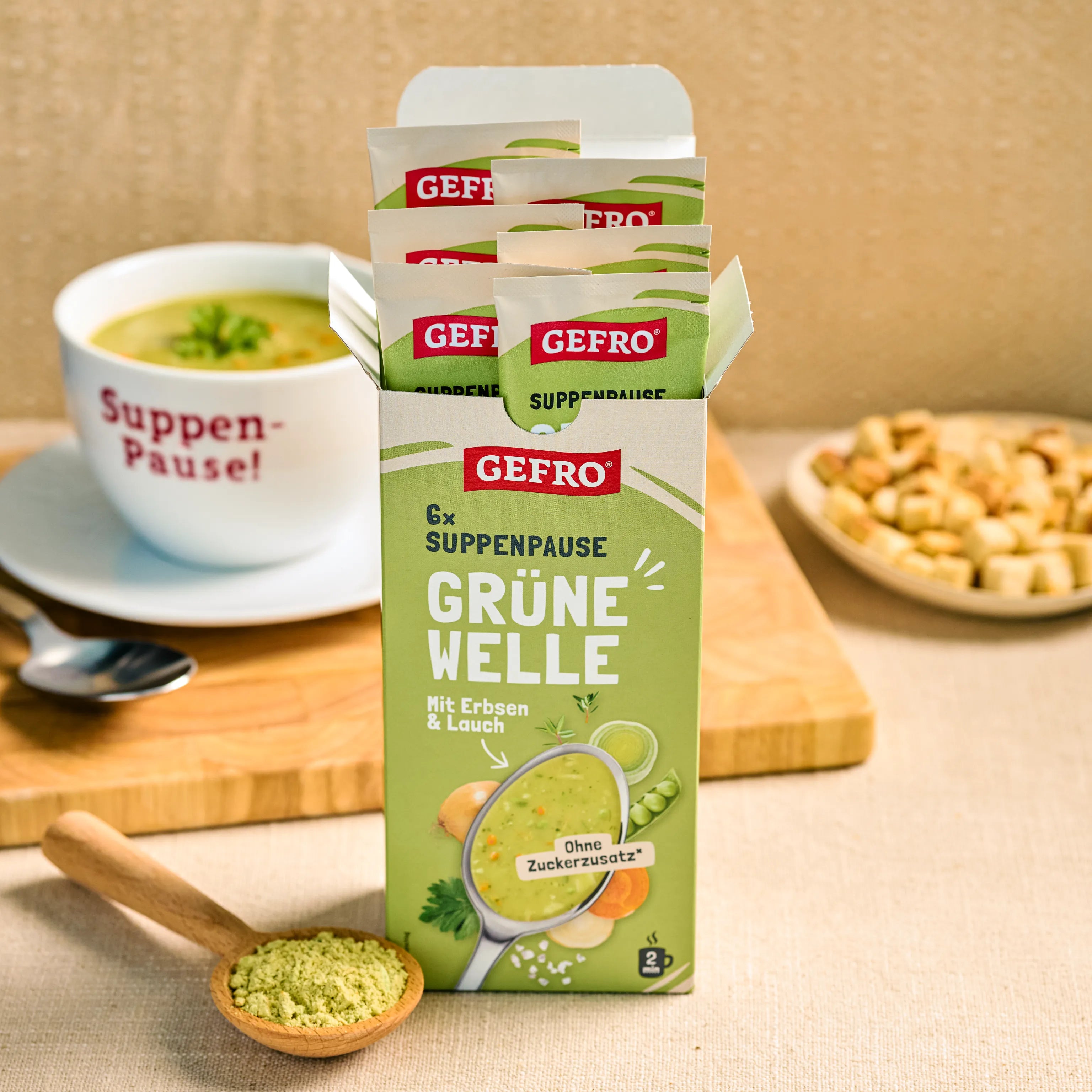 Geöffnete Packung GEFRO Suppenpause Grüne Welle mit Portionsbeuteln, davor Pulverlöffel und im Hintergrund eine Tasse Suppe
