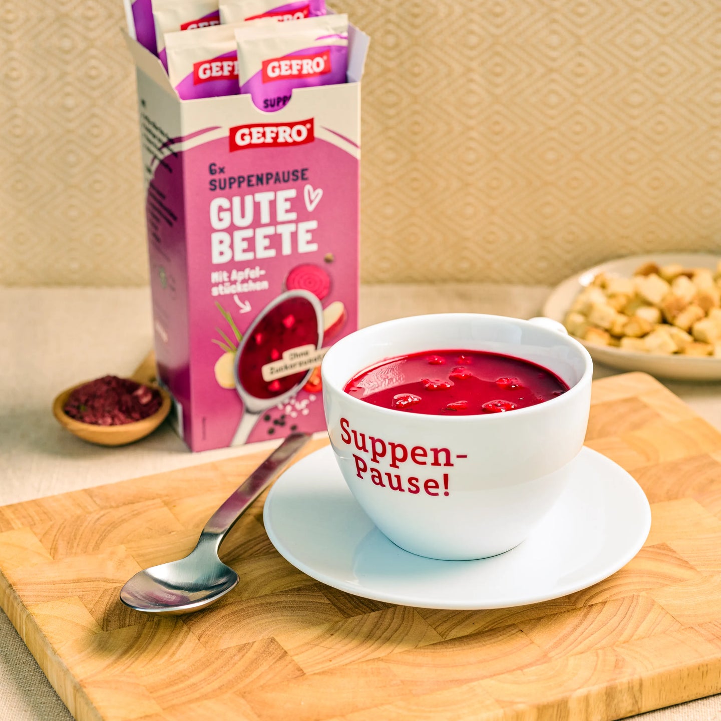 Tasse mit roter Bete-Suppe auf Untertasse neben Löffel, dahinter geöffnete GEFRO-Packung Gute Beete.