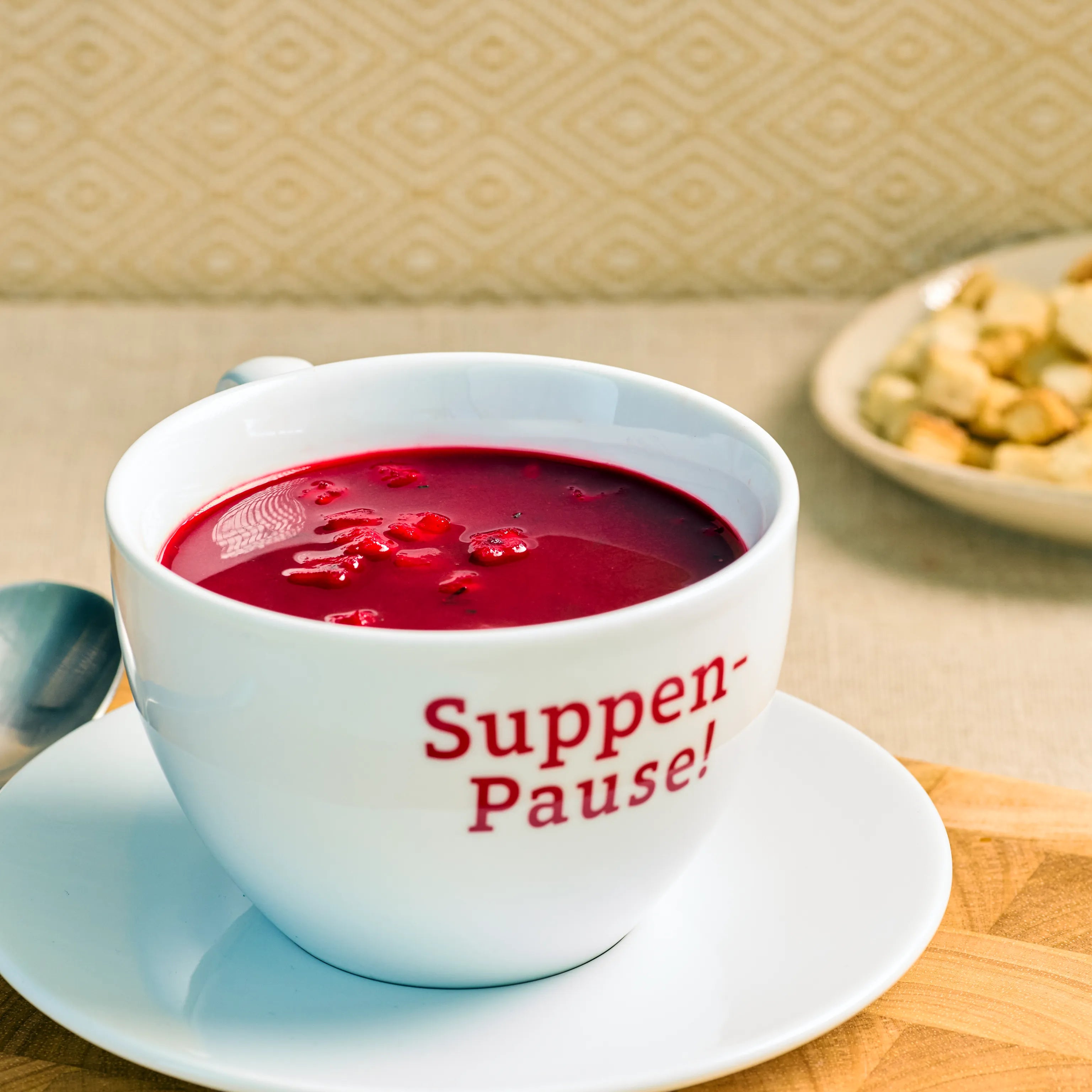 Weiße Suppentasse mit roter Suppe auf Untertasse, daneben Löffel; im Hintergrund steht ein Teller mit Croutons.