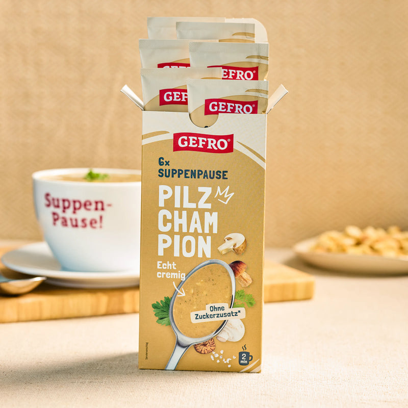 GEFRO Suppenpause Pilz-Champion in geöffneter Packung mit Portionsbeuteln, daneben eine Tasse Suppe im Hintergrund
