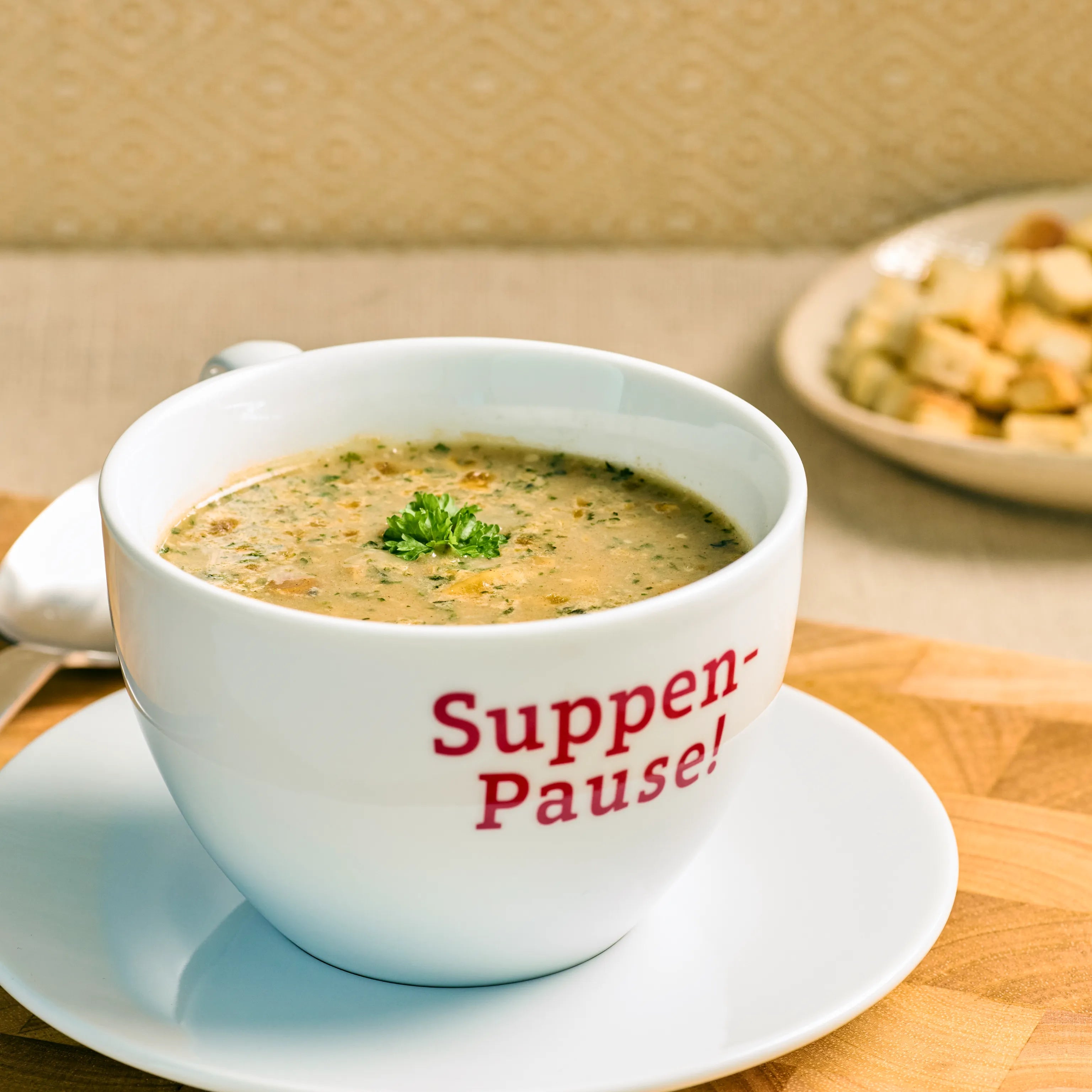 Weiße Tasse mit cremiger Suppe auf Untertasse, garniert mit Petersilie, daneben Löffel und Brotwürfel im Hintergrund