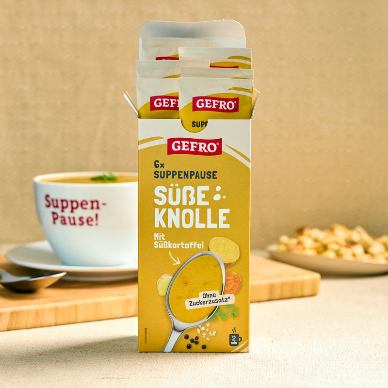 Geöffnete GEFRO Suppenpause Süße Knolle mit Portionsbeuteln, davor eine Tasse Suppe auf Holzbrett.