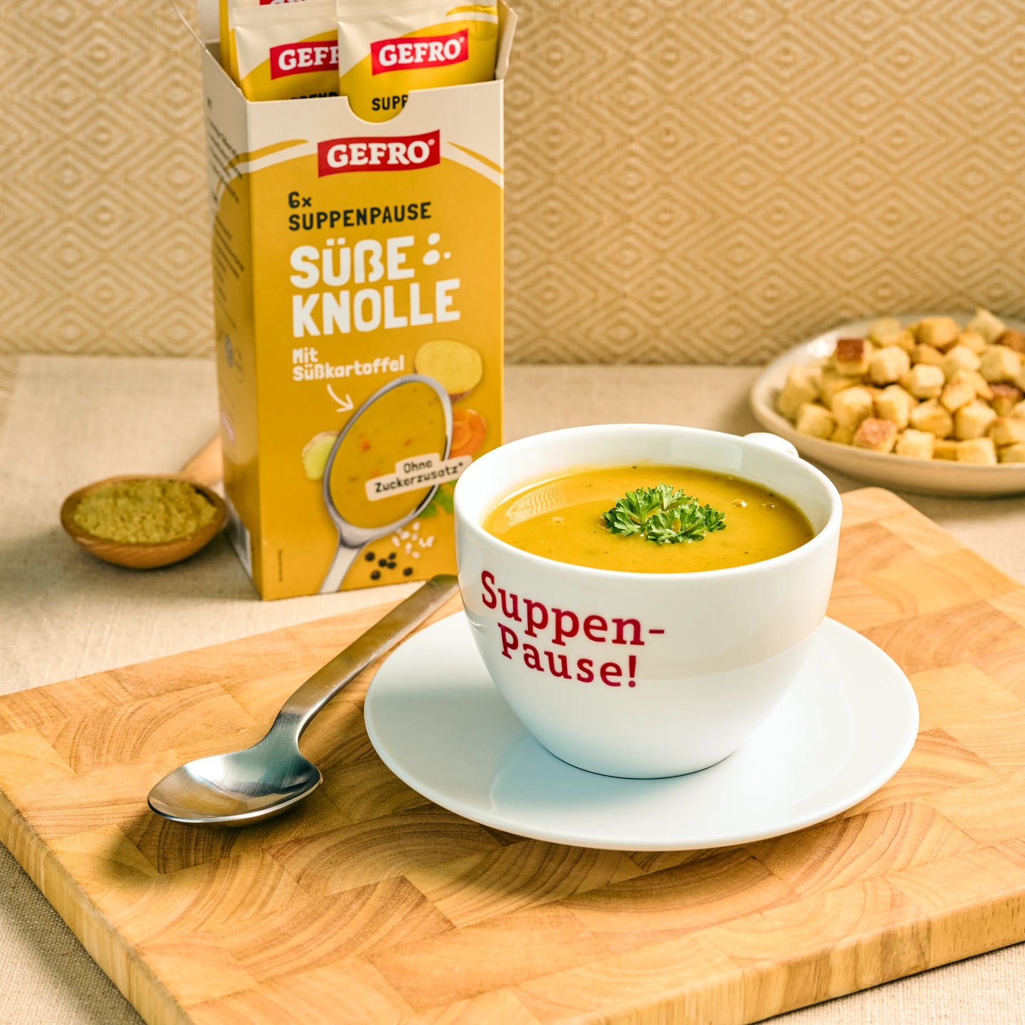 Schale mit cremiger Süßkartoffelsuppe auf Untertasse, daneben Löffel und GEFRO Verpackung Süße Knolle im Hintergrund