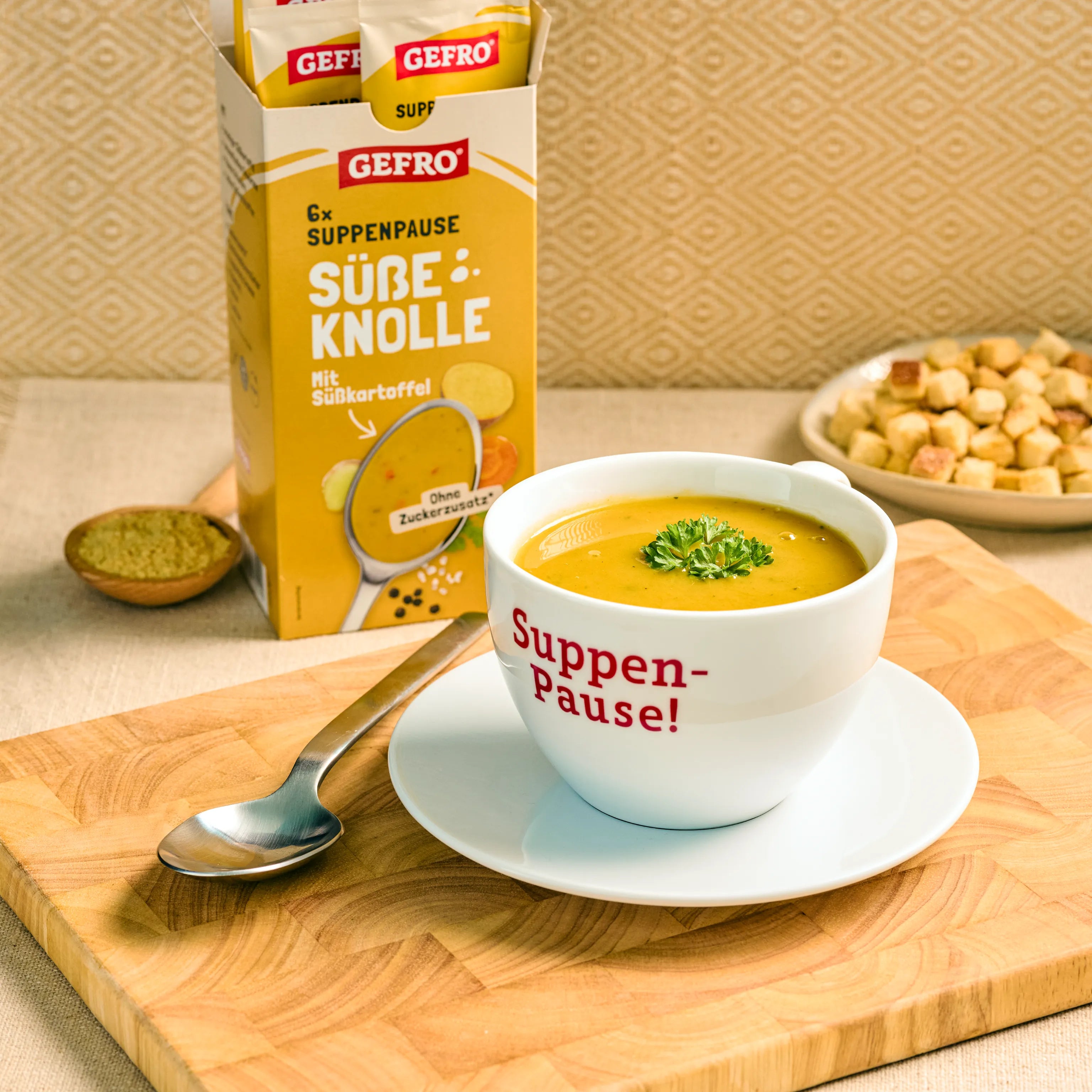 Schale mit cremiger Süßkartoffelsuppe auf Untertasse, daneben Löffel und GEFRO Verpackung Süße Knolle im Hintergrund