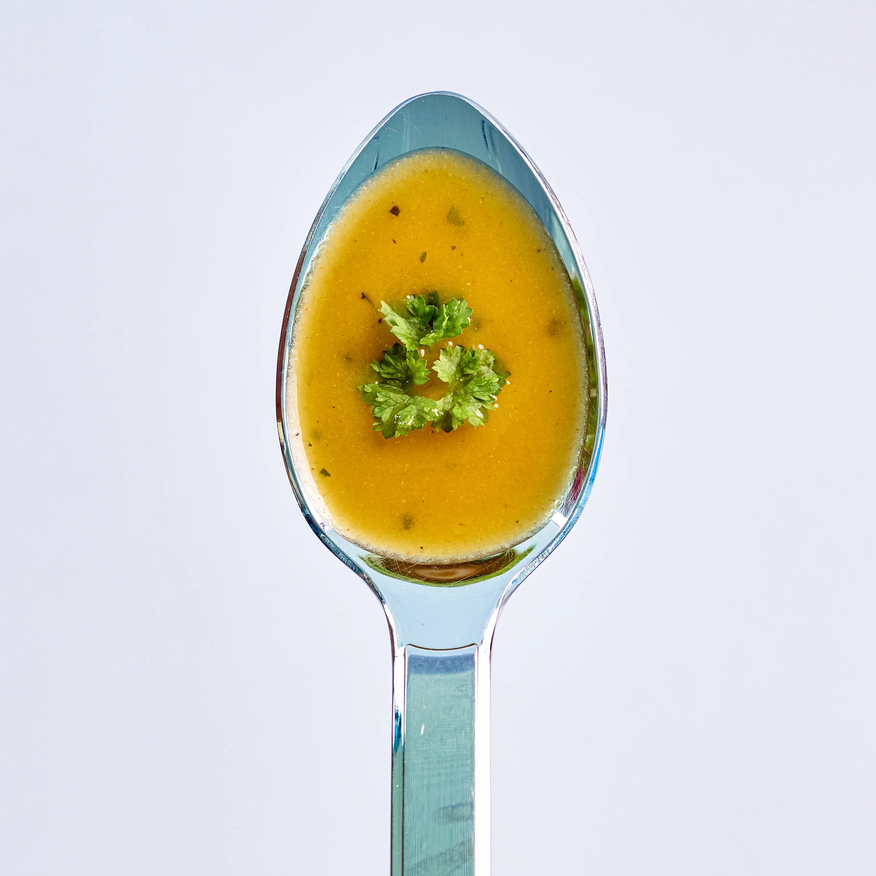 Löffel mit gelber Cremesuppe, von oben fotografiert, mit Petersilie garniert vor hellem Hintergrund.