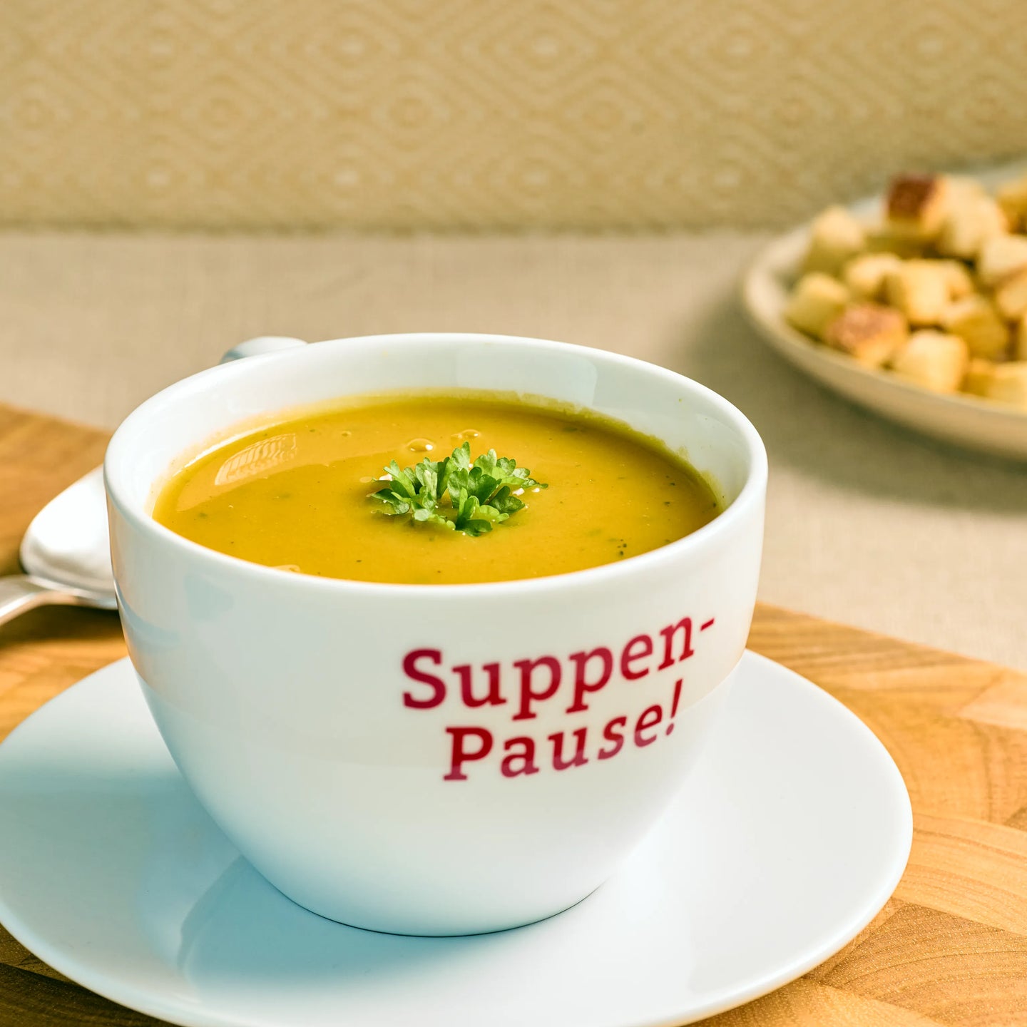 Weiße Tasse mit gelber Cremesuppe und Petersilie auf Unterteller, Aufdruck „Suppen-Pause!“ auf der Vorderseite