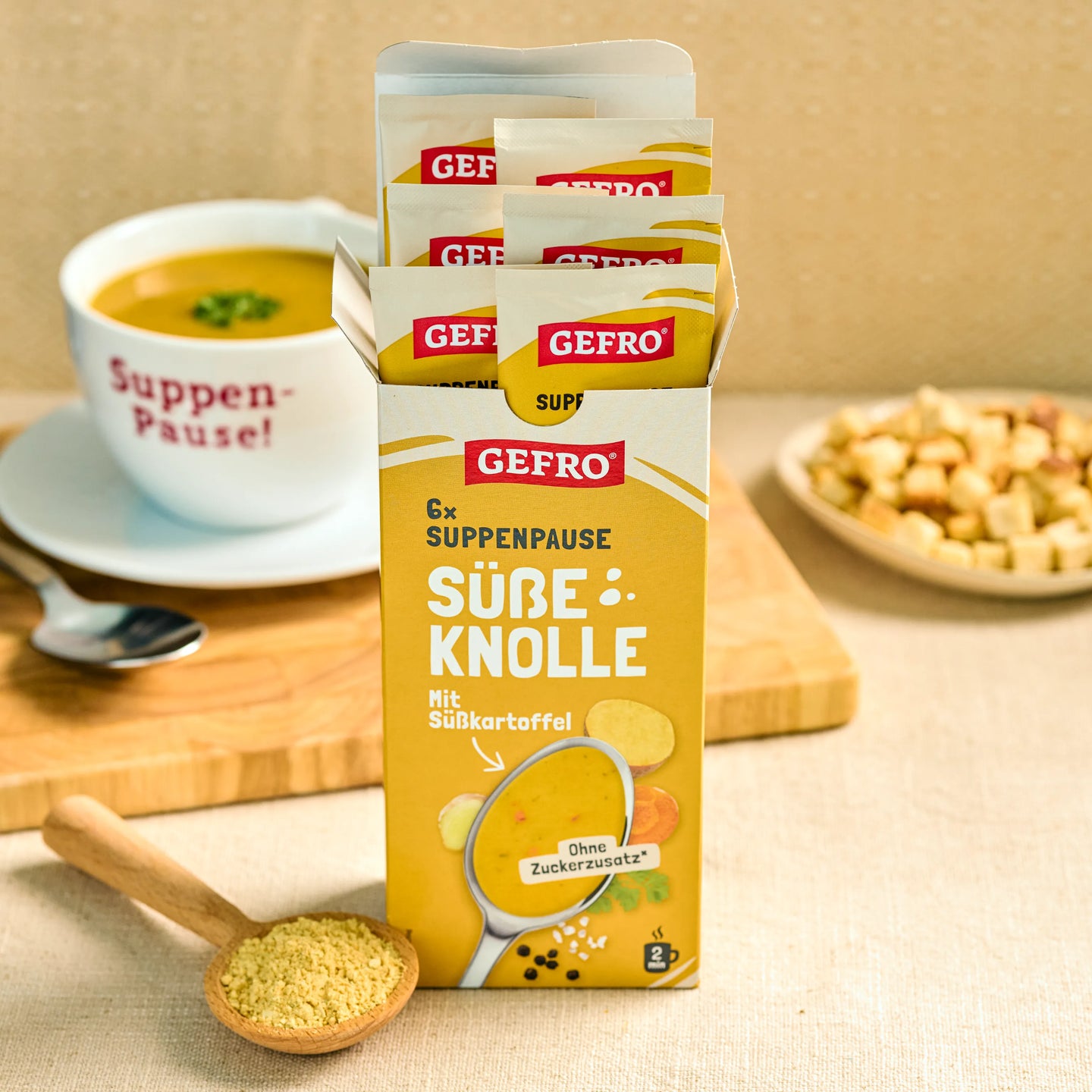 GEFRO Suppenpause Süße Knolle in Portionsbeuteln, davor Pulver im Holzlöffel und eine Tasse Suppe im Hintergrund