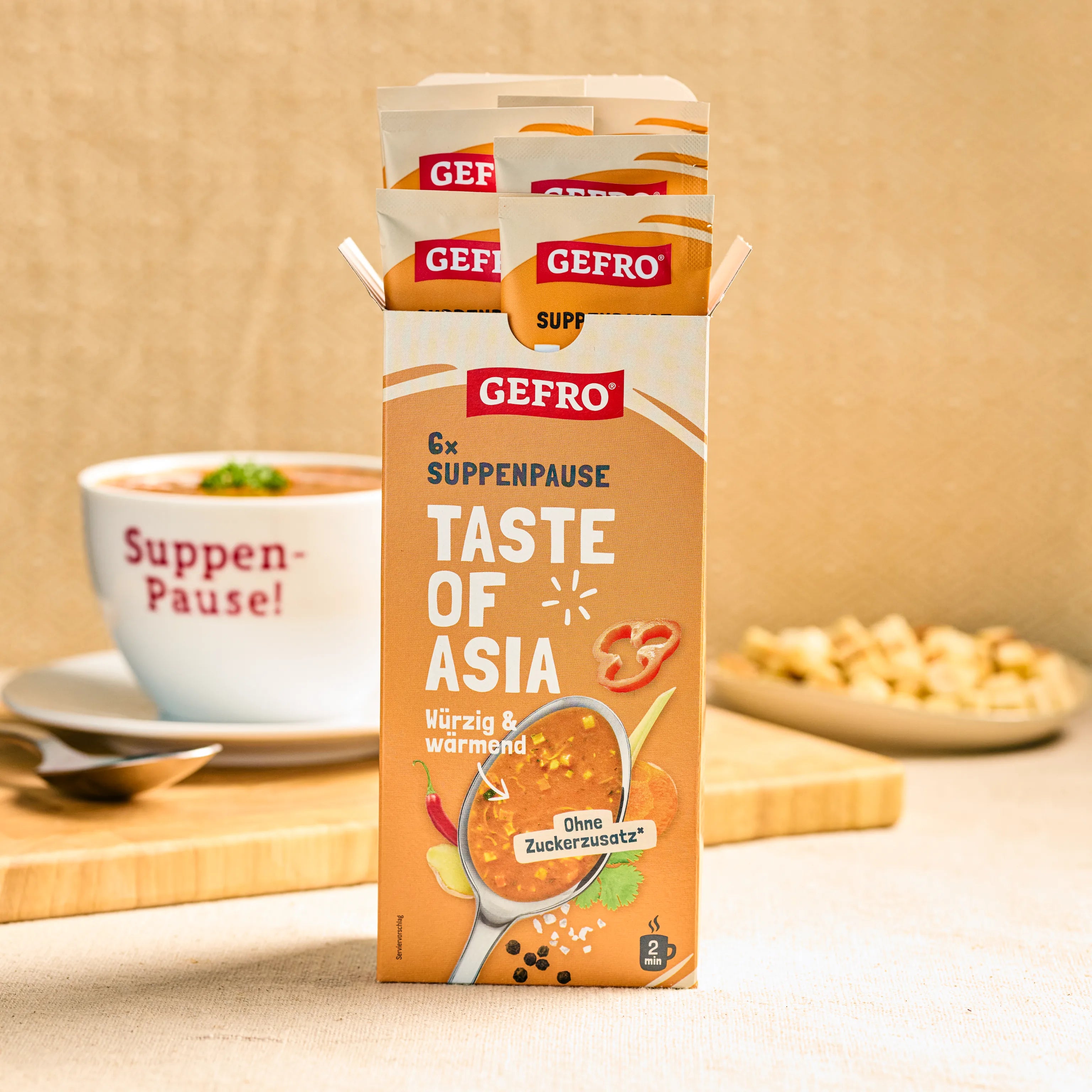 Geöffnete GEFRO Suppenpause Taste of Asia Packung mit Portionsbeuteln, daneben eine Tasse Suppe im Hintergrund
