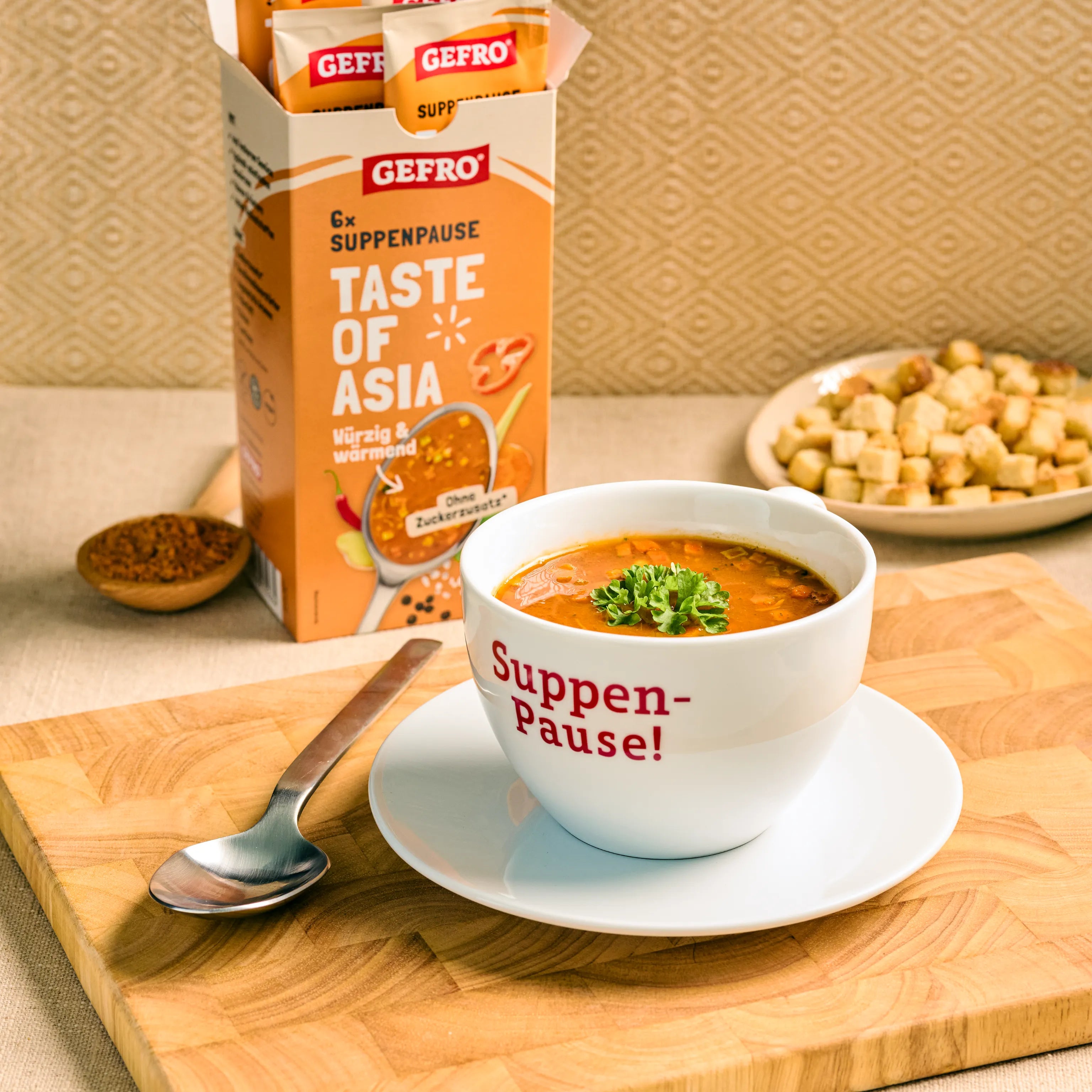 Tasse mit asiatischer Suppe auf Unterteller vor GEFRO Suppenpause Taste of Asia Verpackung und Croûtons im Hintergrund