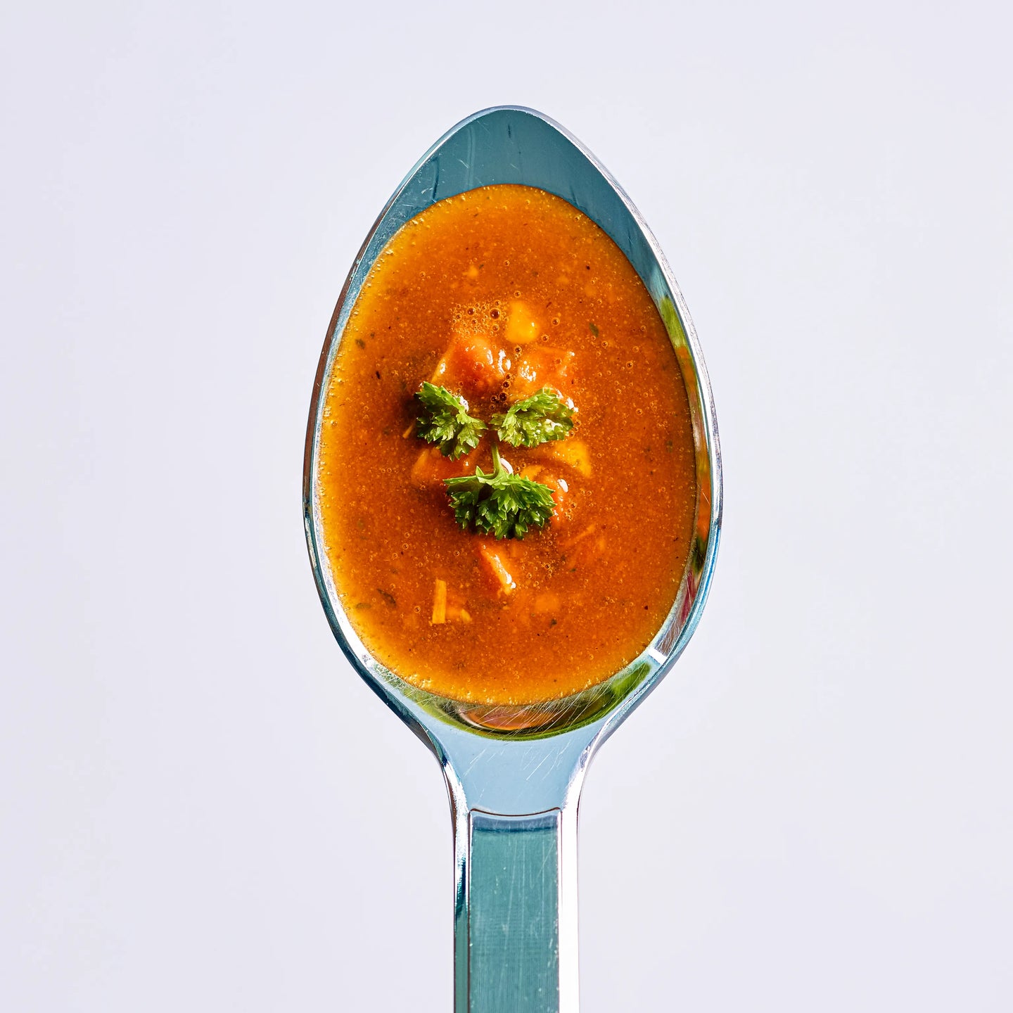Löffel mit roter Suppe und kleinen Gemüsestücken, oben mit Petersilie garniert, auf hellem Hintergrund