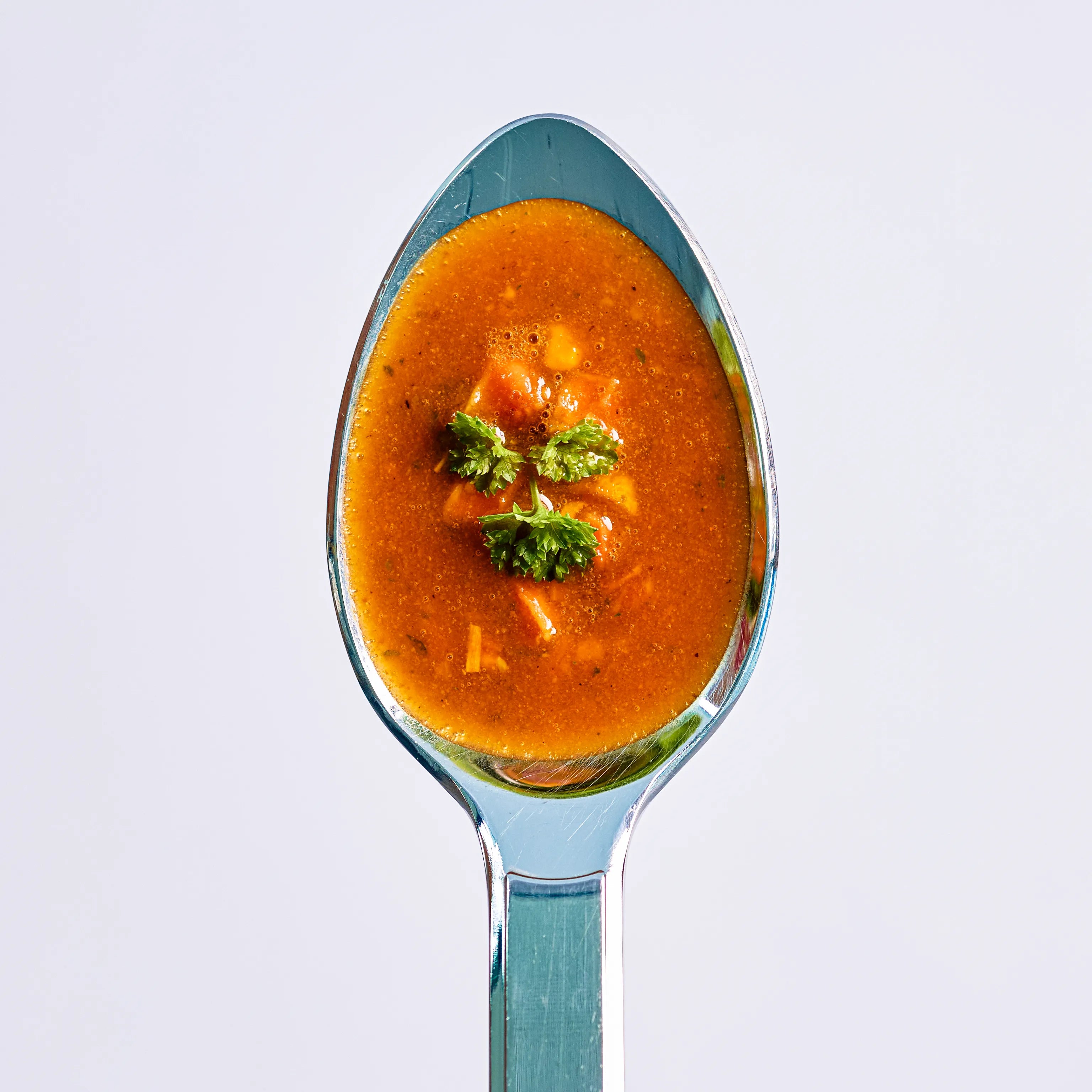 Löffel mit roter Suppe und kleinen Gemüsestücken, oben mit Petersilie garniert, auf hellem Hintergrund