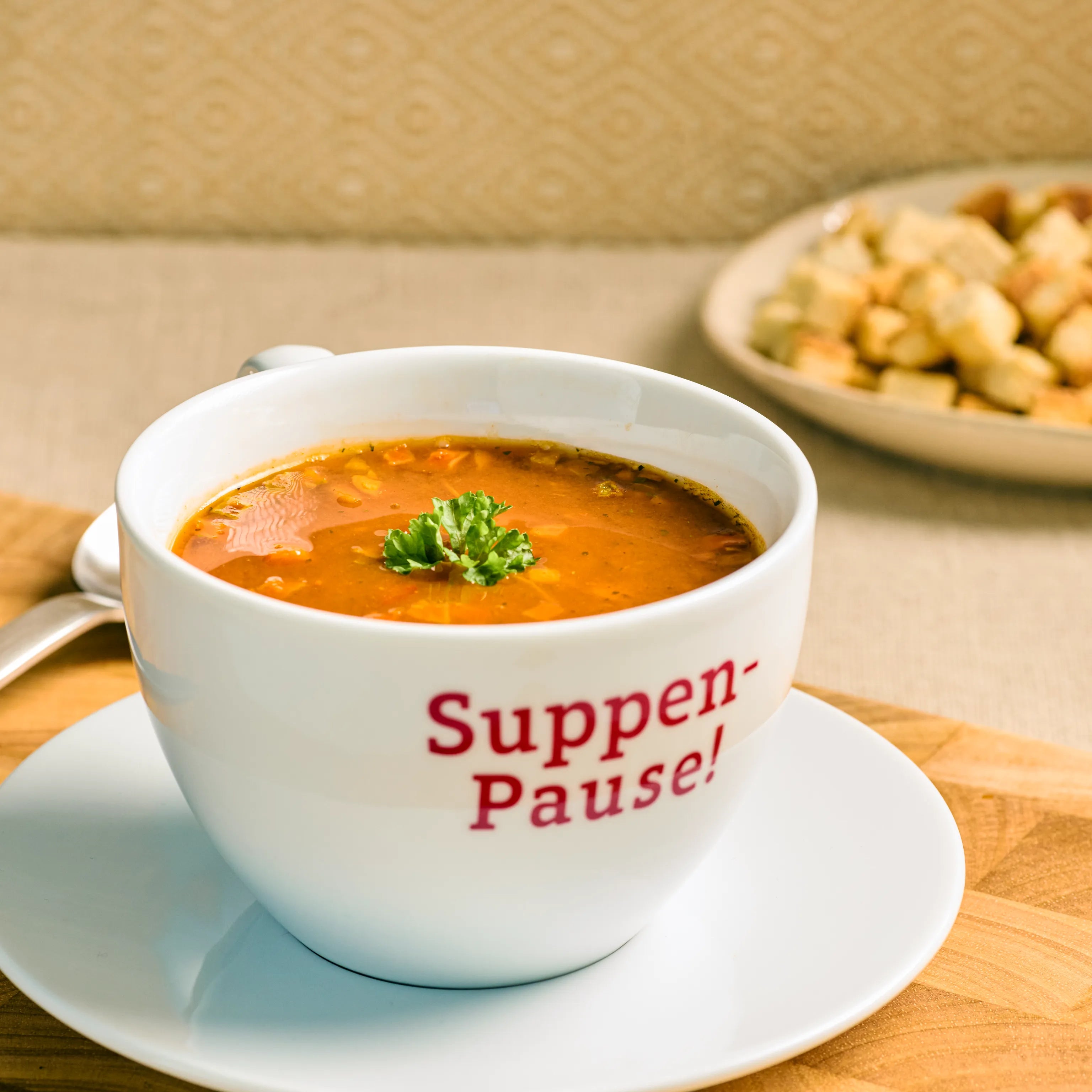 Weiße Tasse mit roter Suppe und Petersilie auf Unterteller, Aufdruck „Suppen-Pause!“, Brotwürfel im Hintergrund