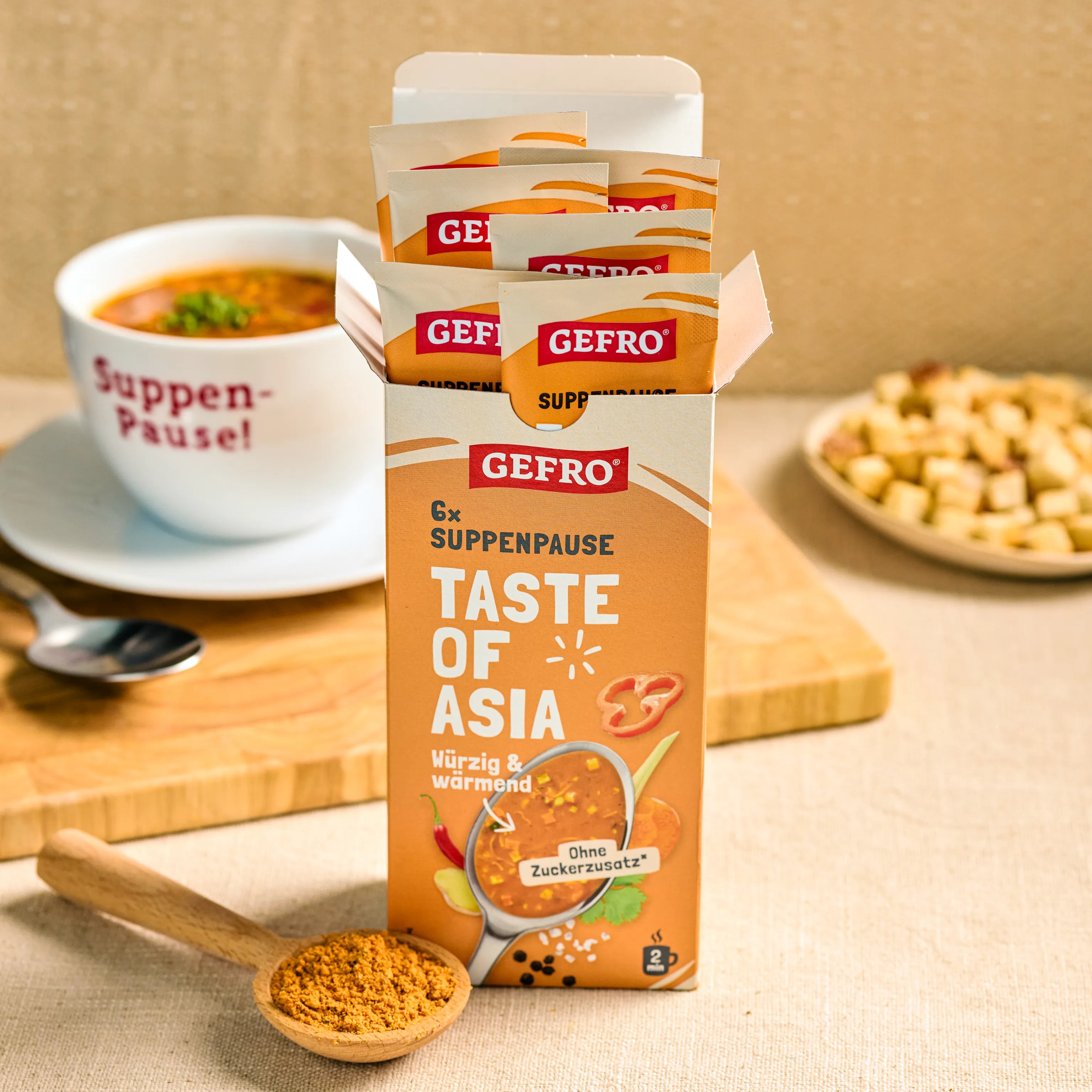 Offene Packung GEFRO Suppenpause Taste of Asia mit Portionsbeuteln, davor Gewürzlöffel und im Hintergrund eine Suppenschale