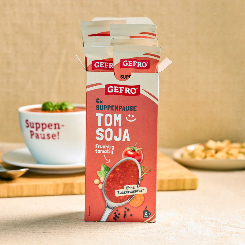 Geöffnete Packung GEFRO Suppenpause Tom Soja mit Portionsbeuteln, davor eine Tasse Tomatensuppe auf dem Tisch