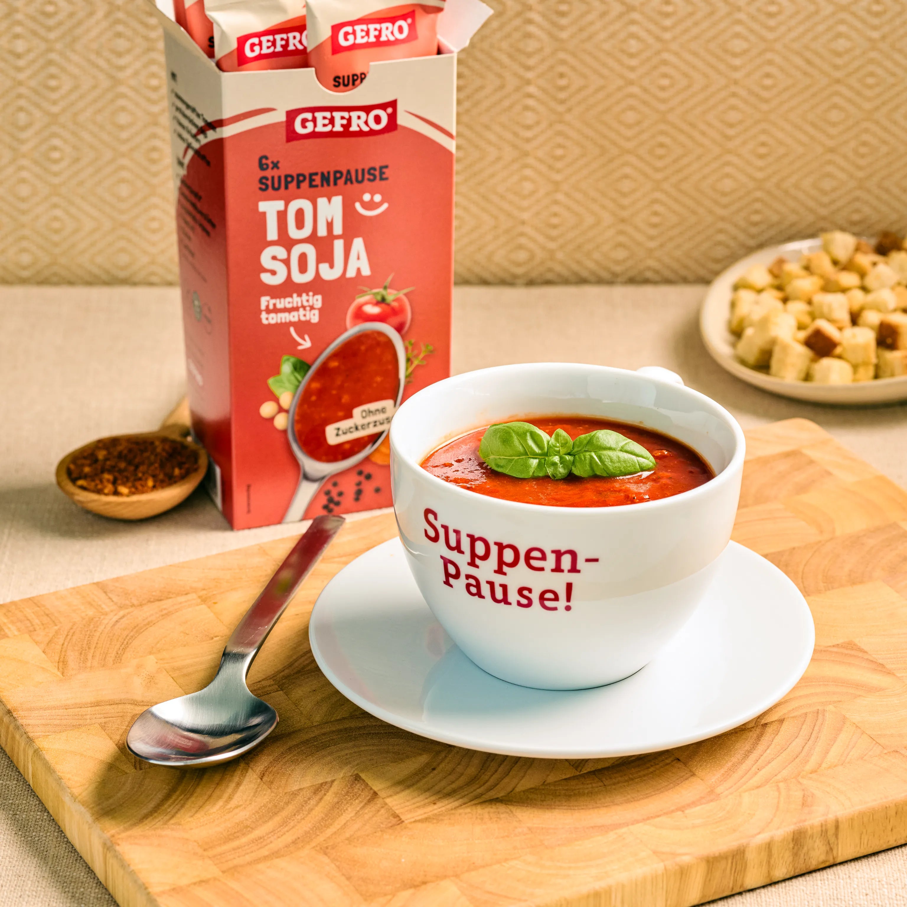 Tasse mit roter Tomaten-Soja-Suppe und Basilikum vor GEFRO Suppenpause Tom Soja Verpackung auf Holzbrett.