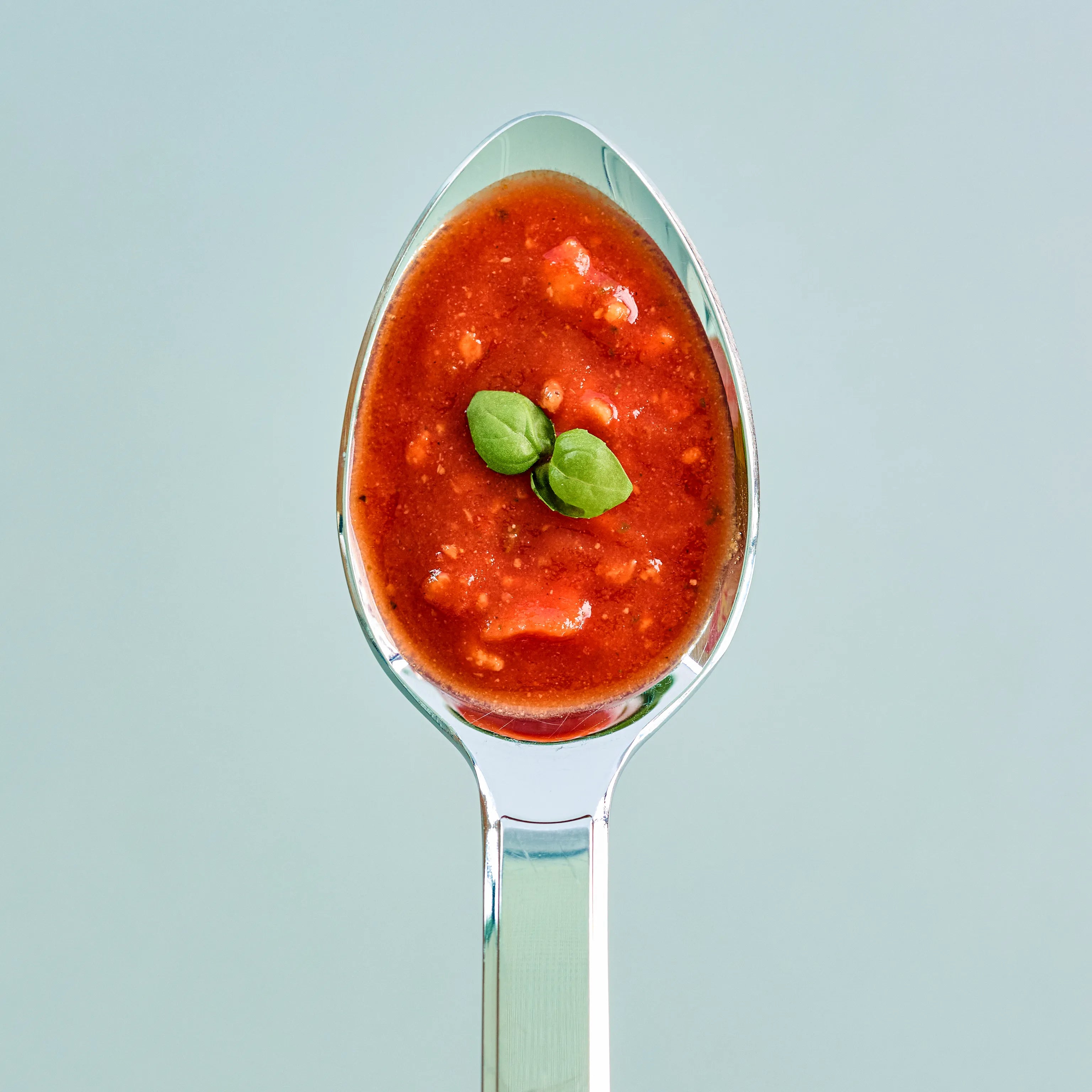 Löffel mit roter Tomatensauce und Basilikumblatt, nah aufgenommen vor hellem Hintergrund zur ansprechenden Präsentation