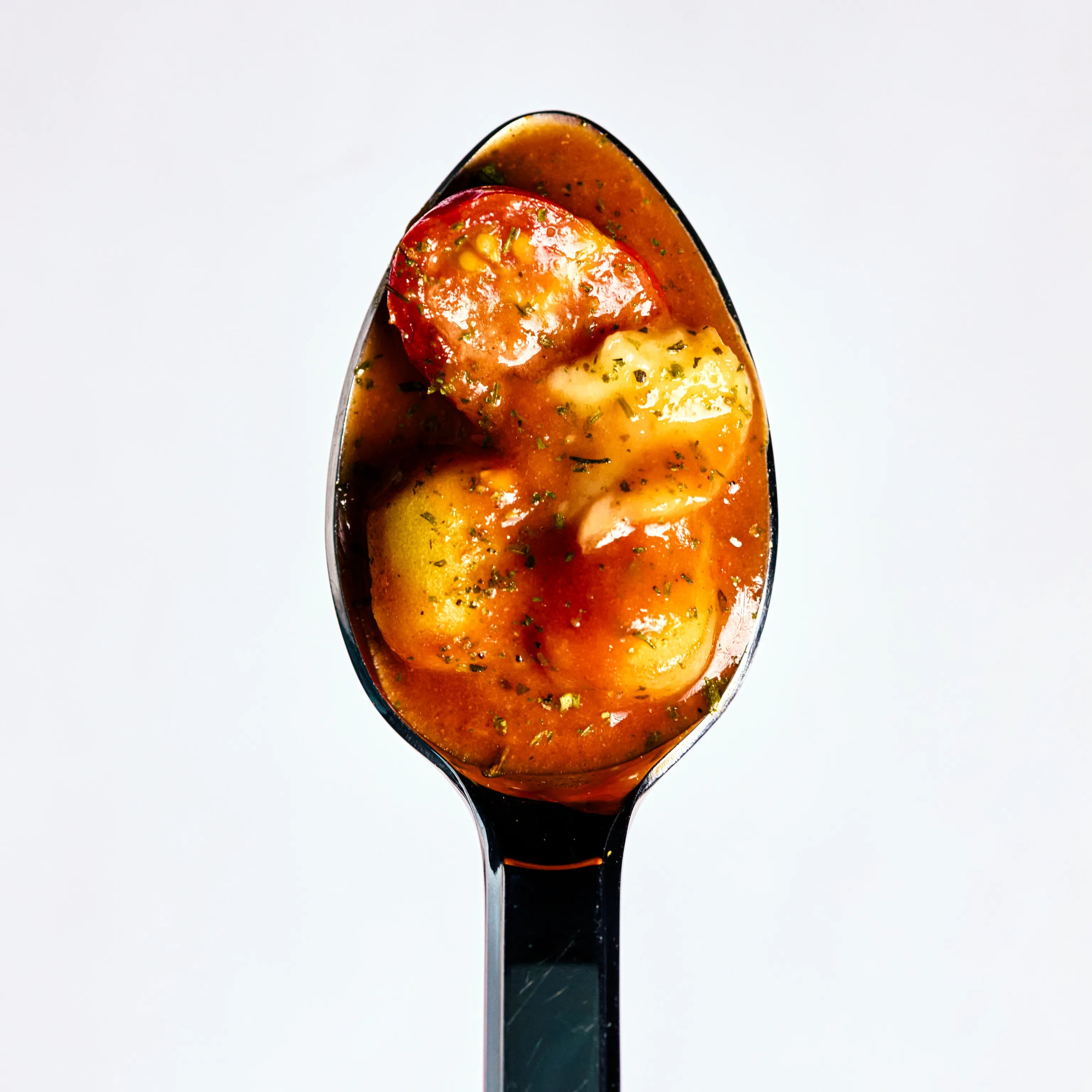 Löffel mit Gnocchi in roter Tomatensauce und einer Scheibe Wurst, nah aufgenommen vor hellem Hintergrund