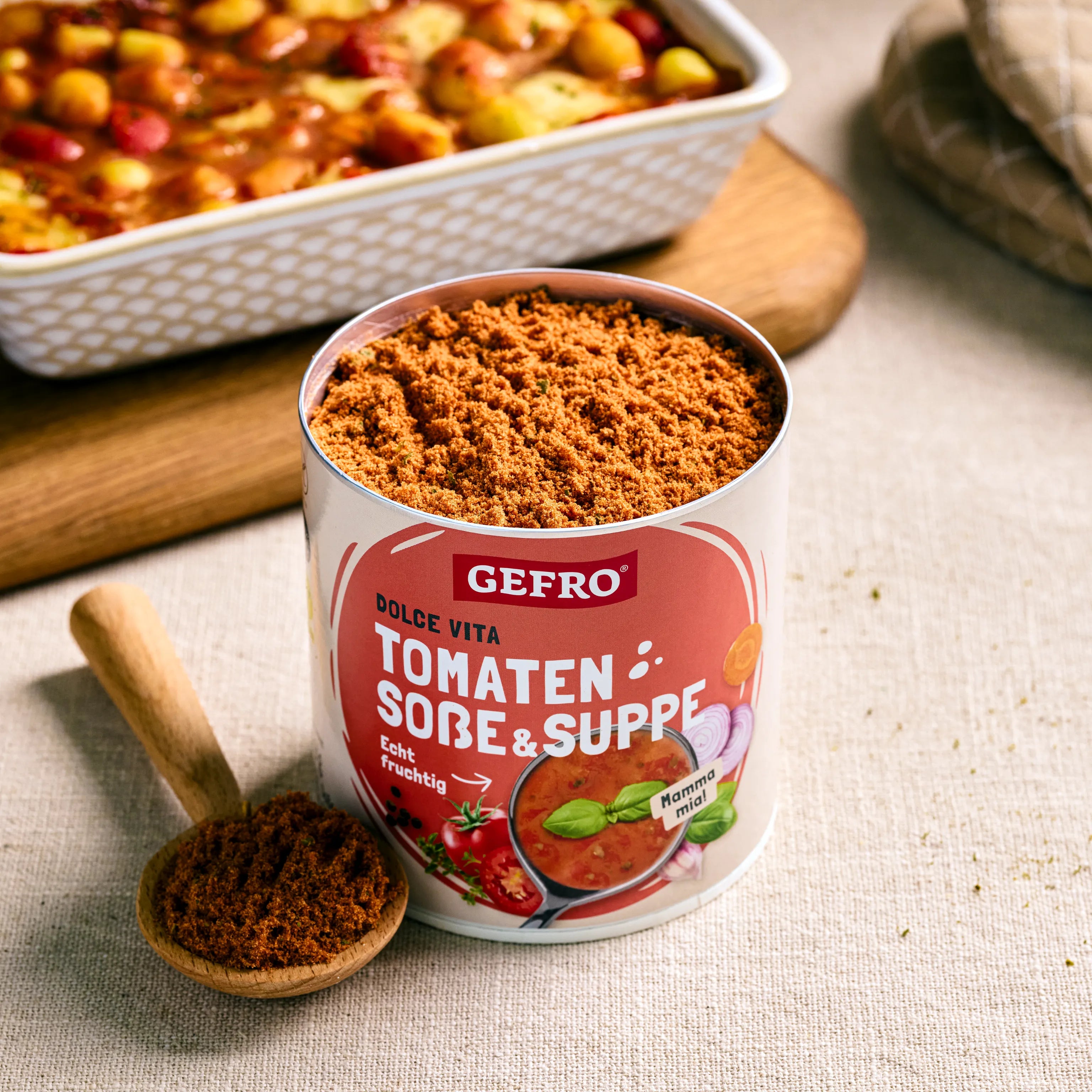 Dose mit GEFRO Tomaten Soße & Suppe Pulver und Holzlöffel, daneben ein Ofengericht im Hintergrund
