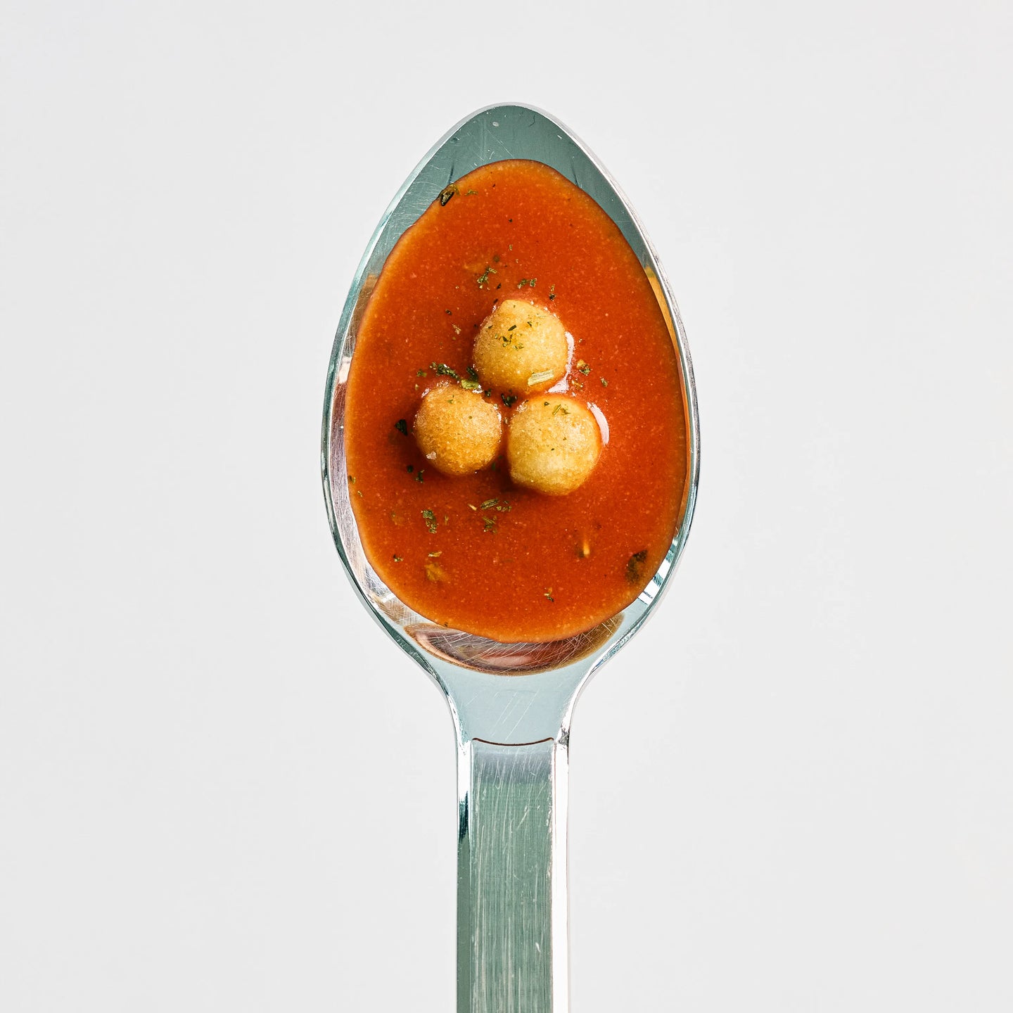 Löffel mit roter Tomatensuppe und drei kleinen runden Einlagen, von oben vor hellem Hintergrund fotografiert