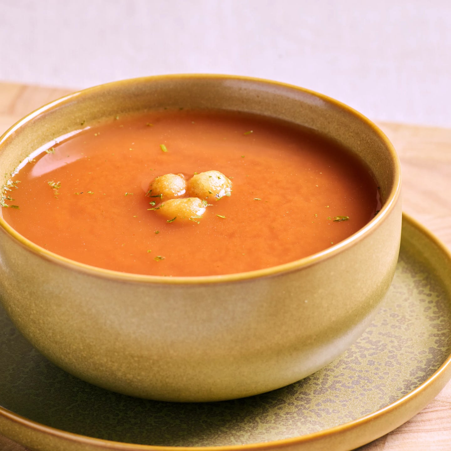 Schale mit cremiger Tomatensuppe auf Unterteller, garniert mit kleinen Croutons und Kräutern auf Holztisch