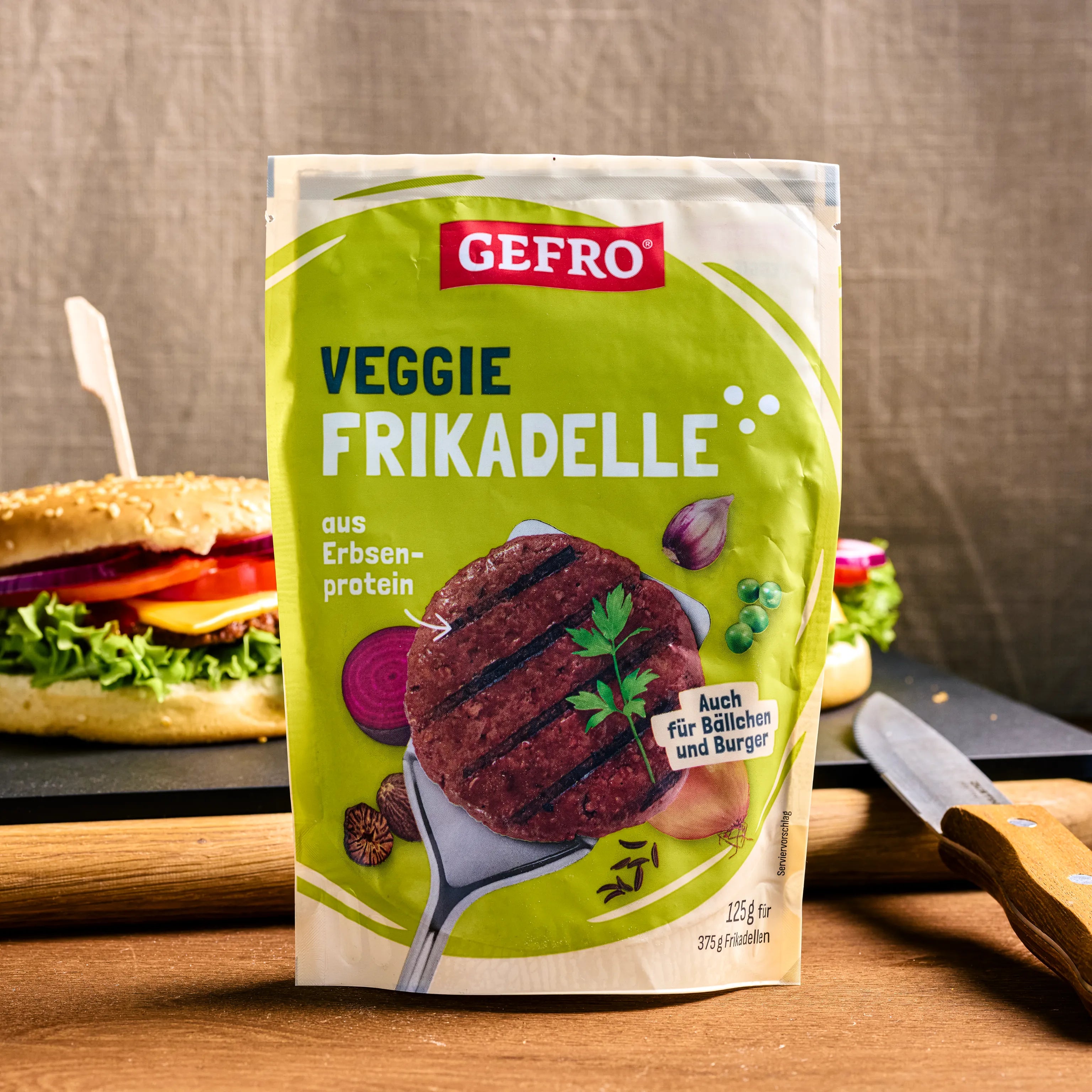 Verpackung von GEFRO Veggie Frikadelle aus Erbsenprotein, aufgestellt vor Burgern und Küchenutensilien.