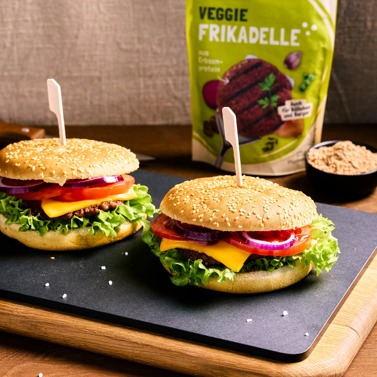Zwei Burger mit Veggie-Frikadelle, Käse, Salat, Tomate und roten Zwiebeln auf Servierplatte, Verpackung im Hintergrund