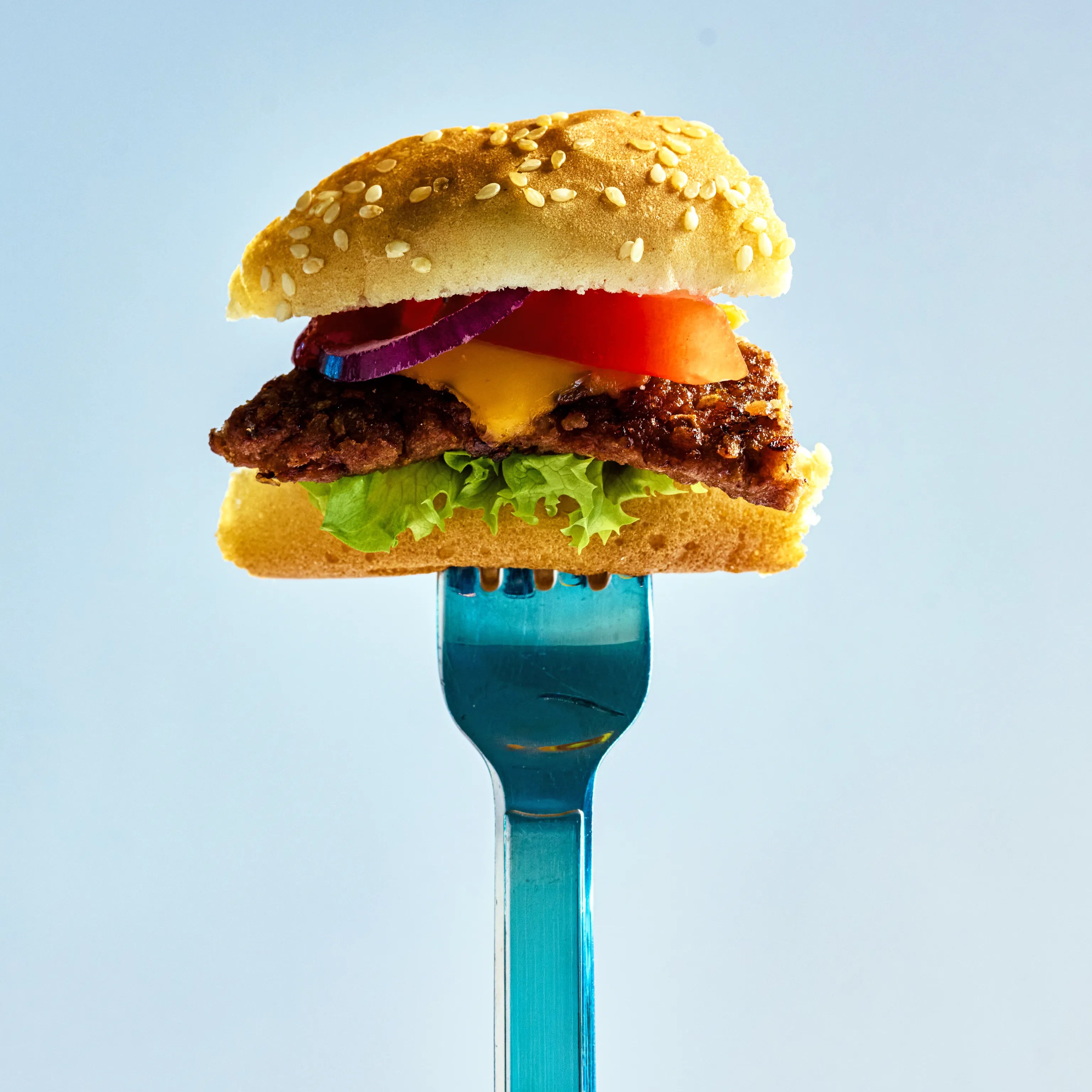 Burger mit Sesambrötchen, Salat, Käse, Tomate und Zwiebeln auf einer blauen Gabel vor hellem Hintergrund