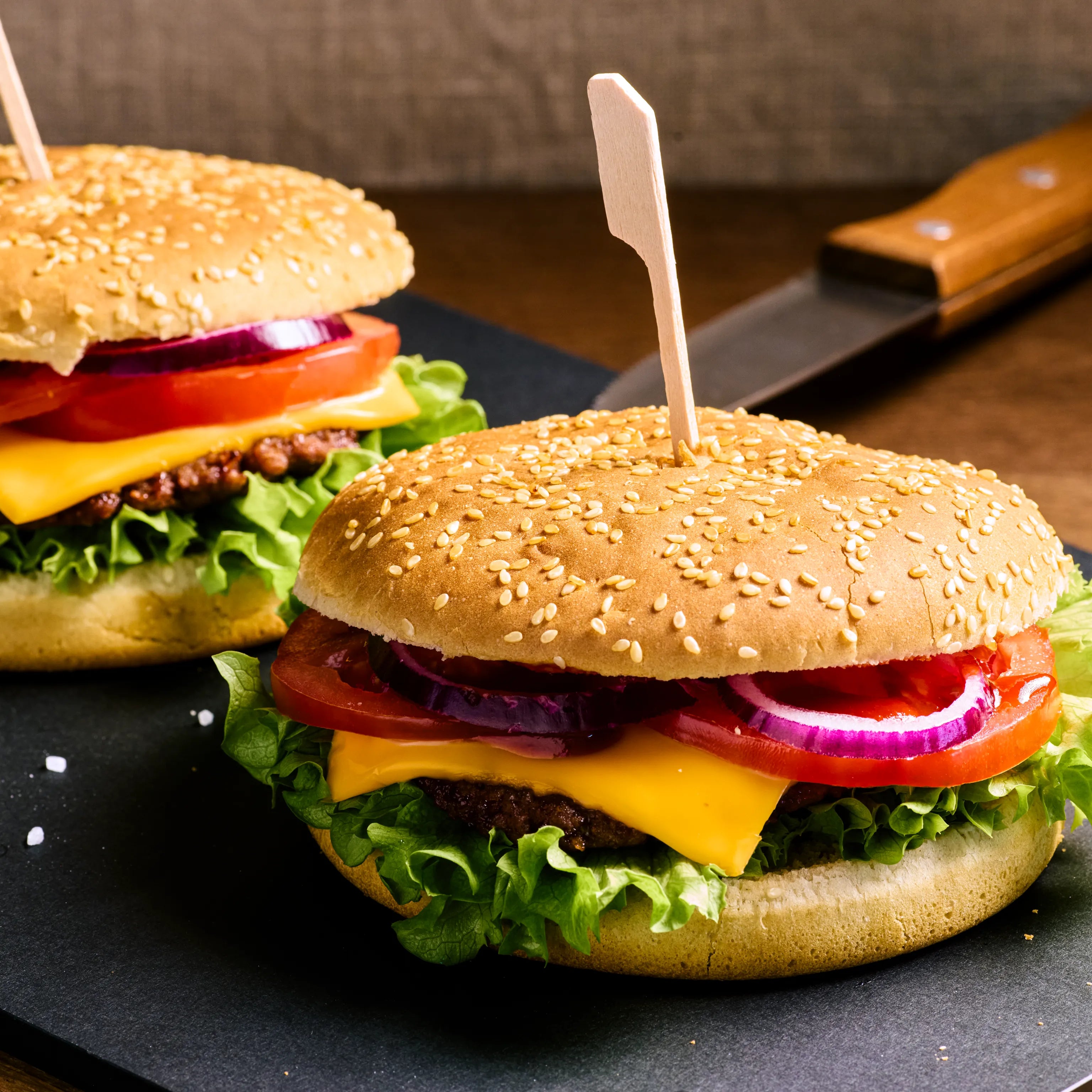 Zwei Cheeseburger mit Sesambrötchen, Salat, Tomaten und roten Zwiebeln auf schwarzer Platte angerichtet.