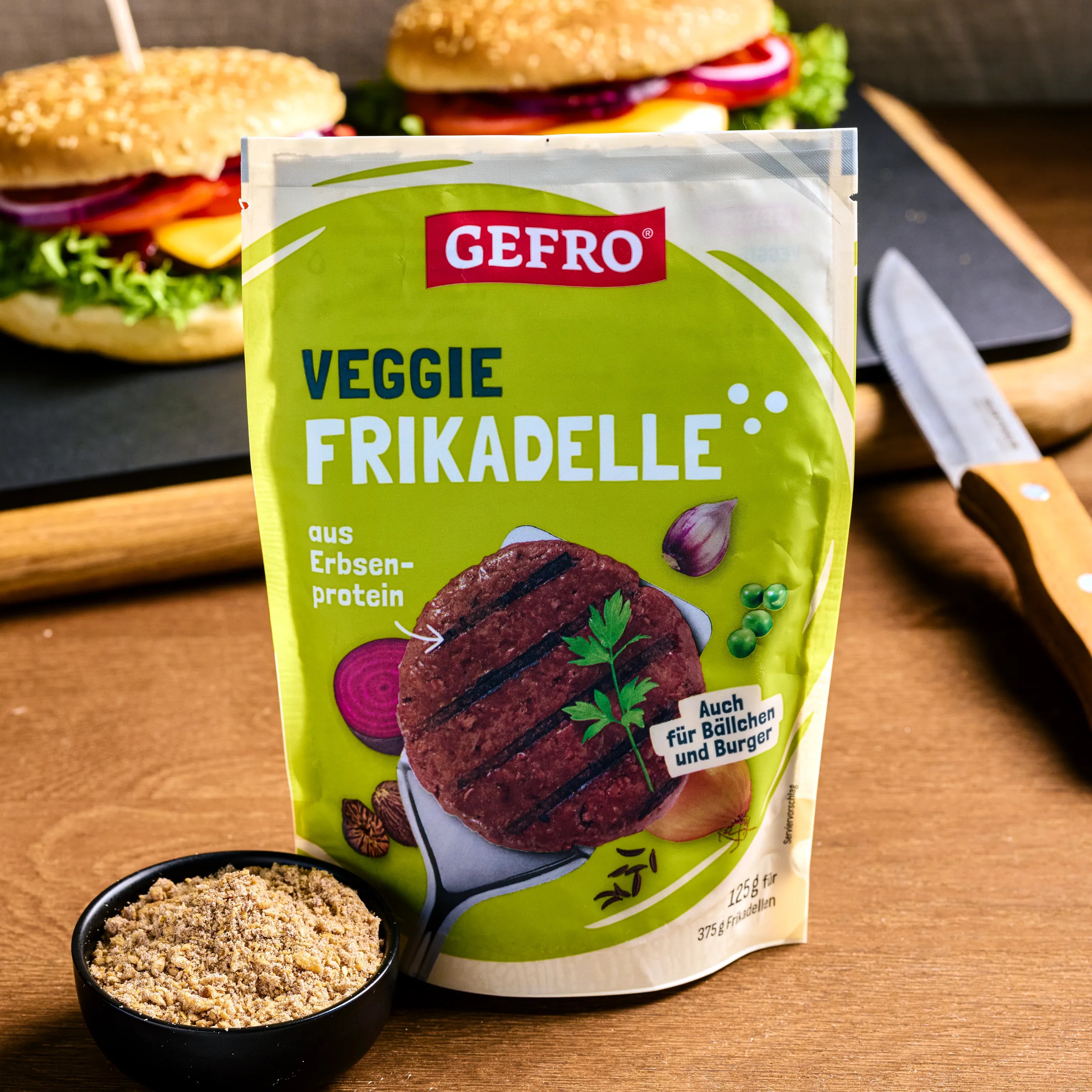 Verpackung der GEFRO Veggie Frikadelle aus Erbsenprotein, davor Pulver in Schale, im Hintergrund zwei Burger