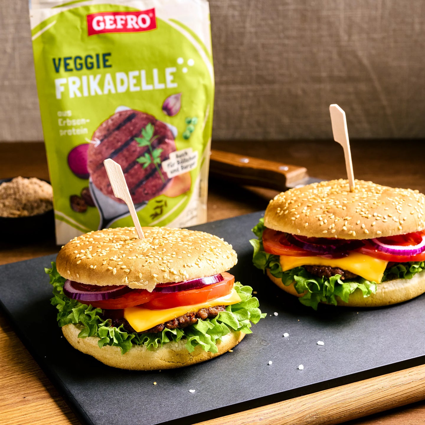 Zwei Burger mit Veggie-Frikadelle, Käse, Salat, Tomate und Zwiebeln auf Platte, dazu Packung im Hintergrund