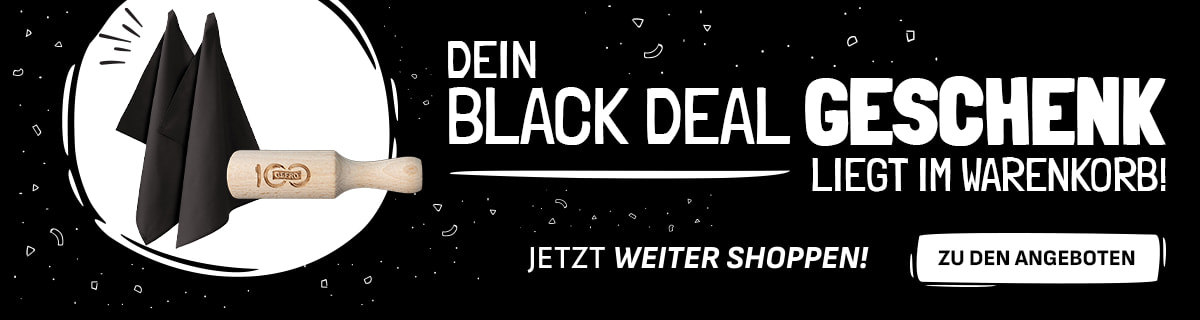 Werbebanner mit schwarzem Hintergrund und weißer Schrift: „Dein Black Deal Geschenk liegt im Warenkorb! Jetzt weiter shoppen!“. Links sind zwei schwarze Stoffservietten und ein hölzernes Nudelholz mit Gravur abgebildet. Rechts befindet sich ein weißer Button mit der Aufschrift „Zu den Angeboten“