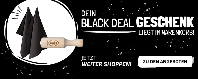 Werbebanner mit schwarzem Hintergrund und weißer Schrift: „Dein Black Deal Geschenk liegt im Warenkorb! Jetzt weiter shoppen!“. Links sind zwei schwarze Stoffservietten und ein hölzernes Nudelholz mit Gravur abgebildet. Rechts befindet sich ein weißer Button mit der Aufschrift „Zu den Angeboten“
